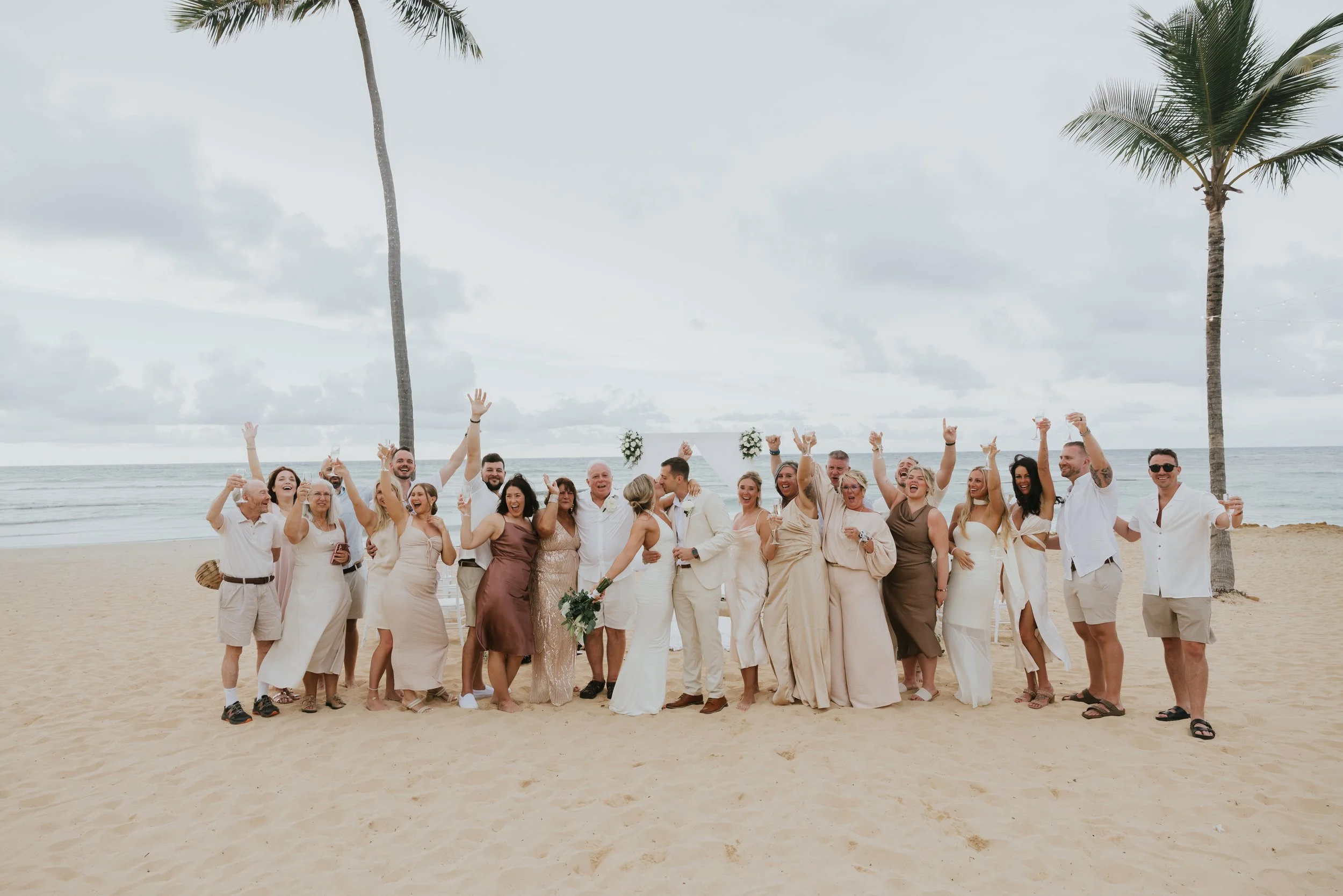 Wedding guests in cream, white and neutral tones cheering with arms raised in a large group photo with the bride and groom on the beach at Secrets Tides, with palm trees and a cloudy sky behind them