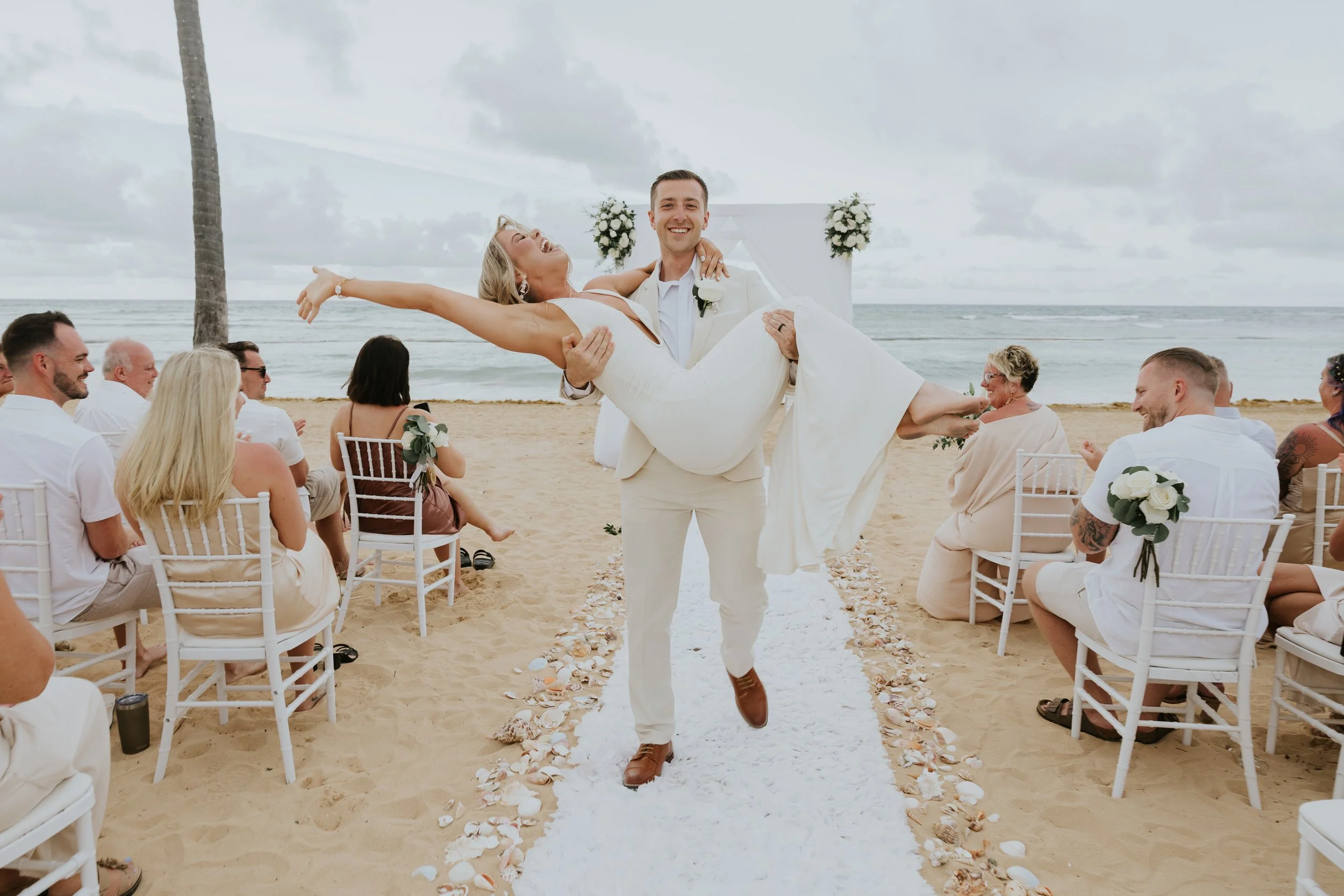 Groom in a cream suit carrying his laughing bride in a white gown down a shell-lined aisle at a Secrets Tides beach ceremony in Punta Cana, with guests seated on white chairs