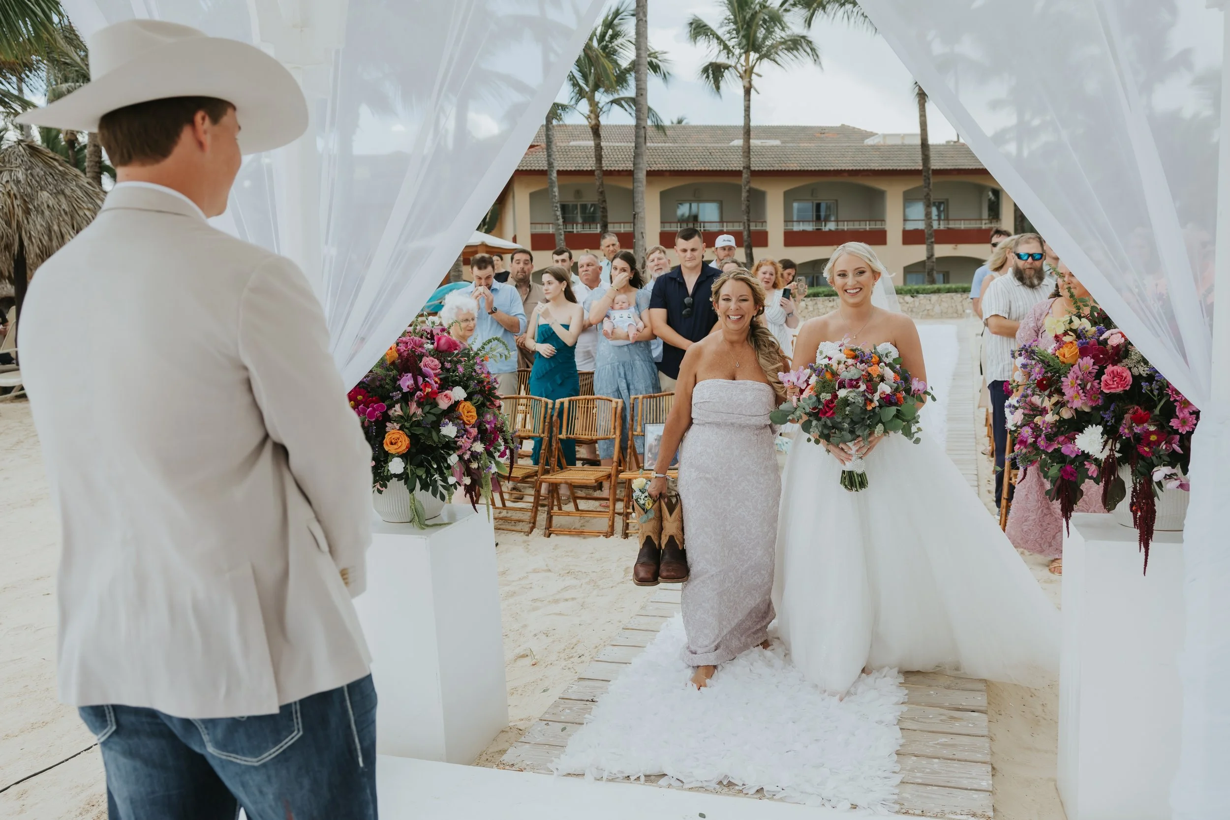 Groom in cowboy hat watching bride and her mother walk the beachfront aisle at their Majestic Elegance Punta Cana wedding ceremony
