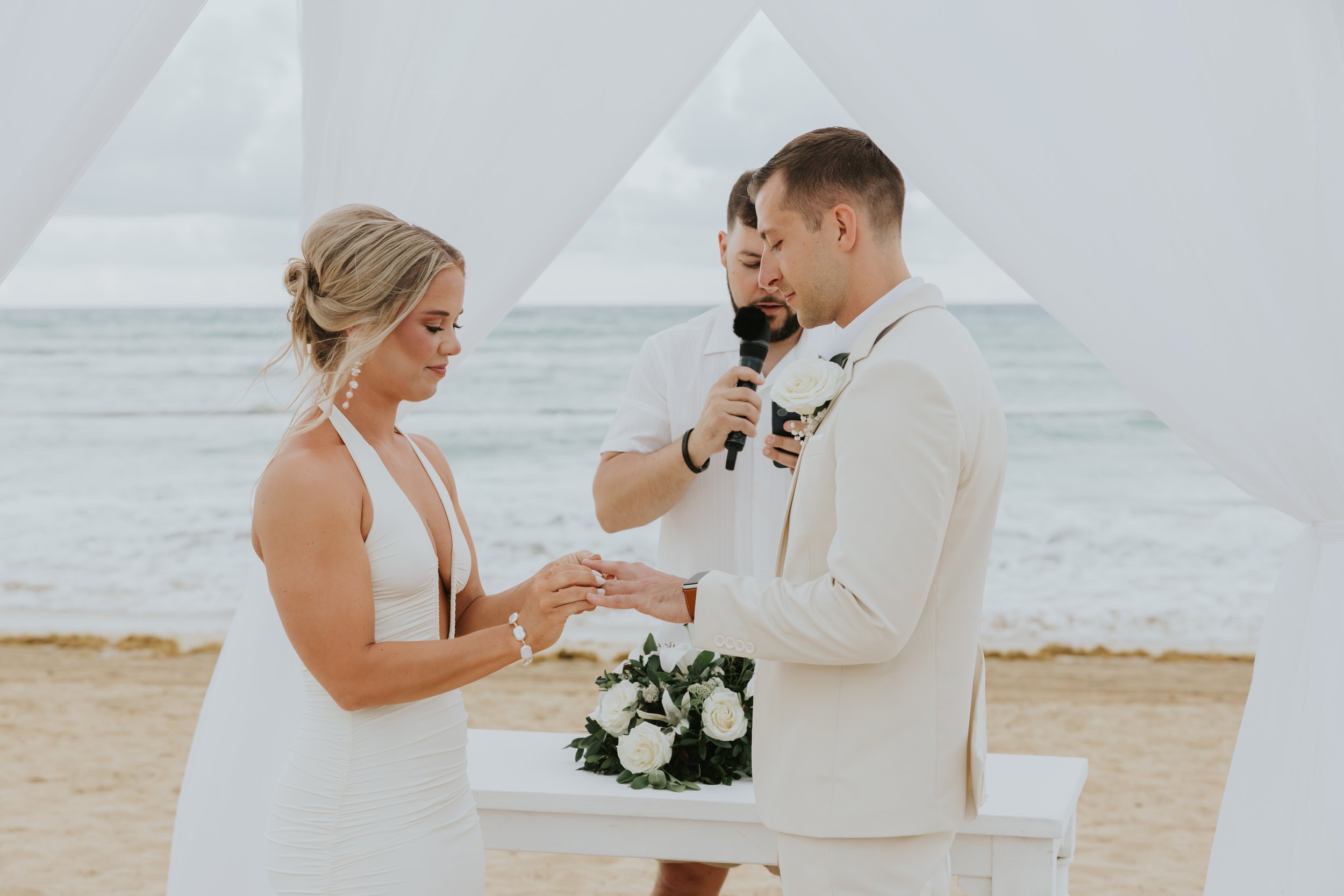 Bride placing a ring on the groom's finger during a beach ceremony at Secrets Tides, while the officiant speaks into a microphone beside a table with white floral arrangements