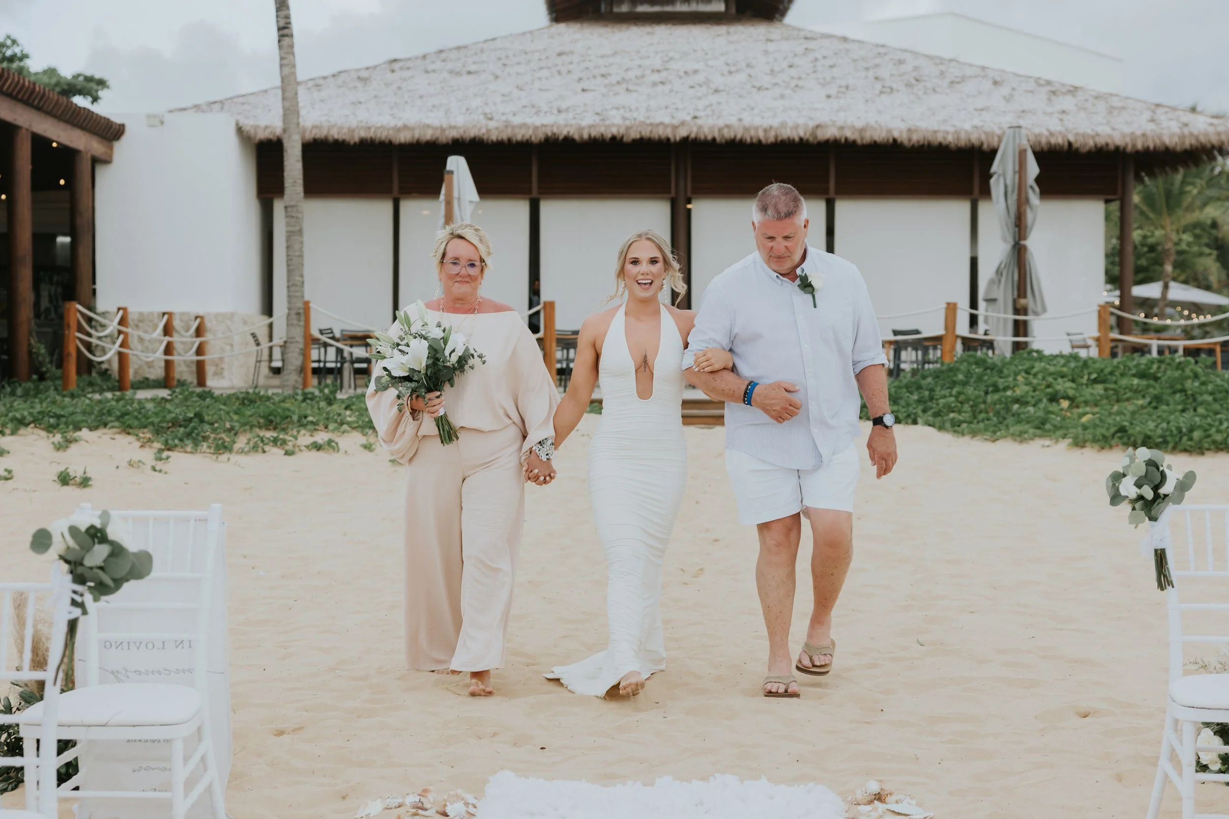Bride in a white plunge-neck gown walking across the sand between her mother and father toward a beach ceremony at Secrets Tides Punta Cana, with the thatched palapa behind them