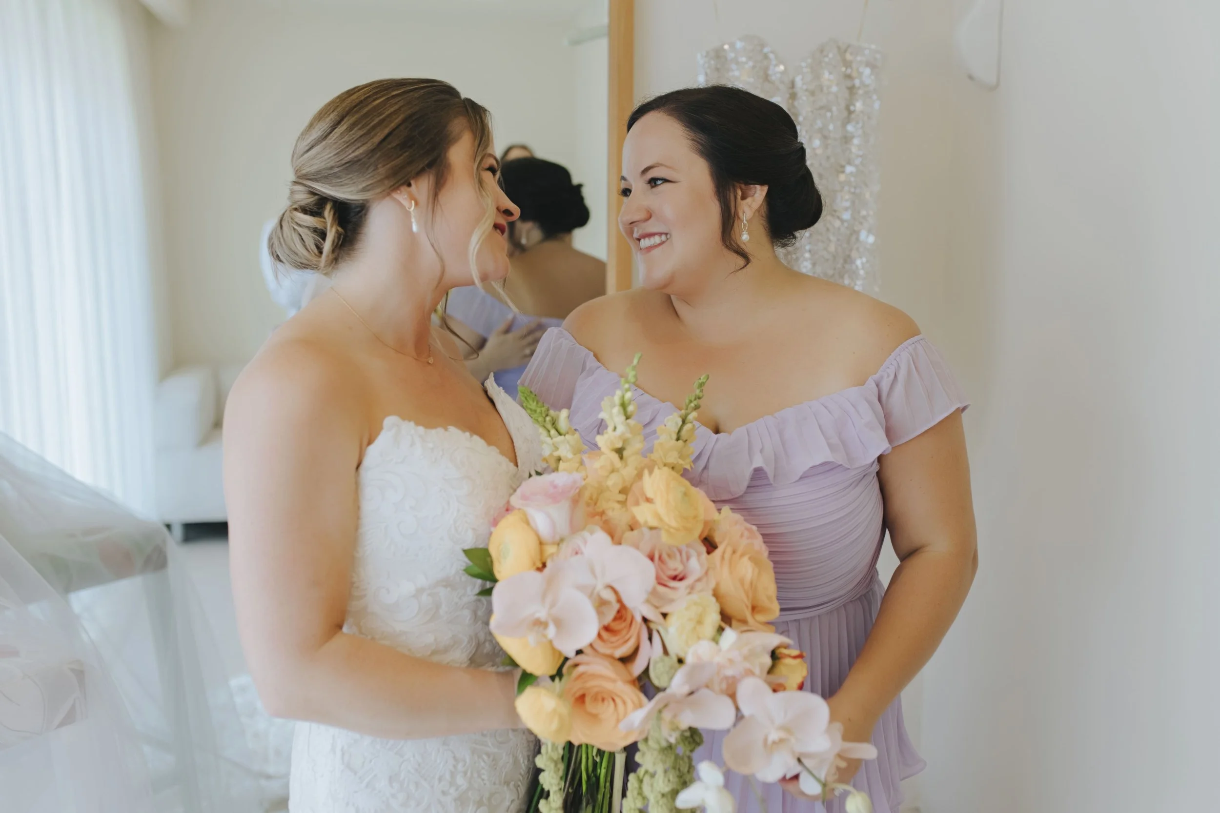 Bride and bridesmaid smiling at each other in the suite before a Punta Cana wedding, both wearing updos and holding peach and blush bouquets