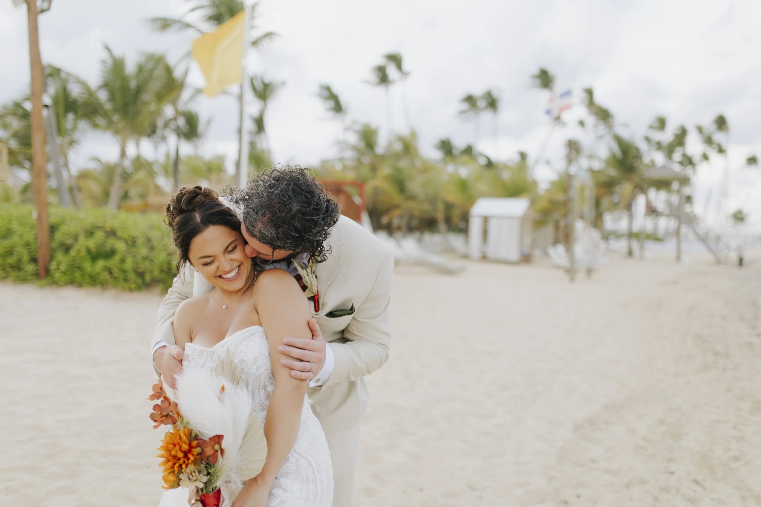 Groom in a cream linen suit hugs his laughing bride from behind and kisses her cheek on a Dreams Onyx beach in Punta Cana, with palm trees, a yellow flag and white beach cabana behind them