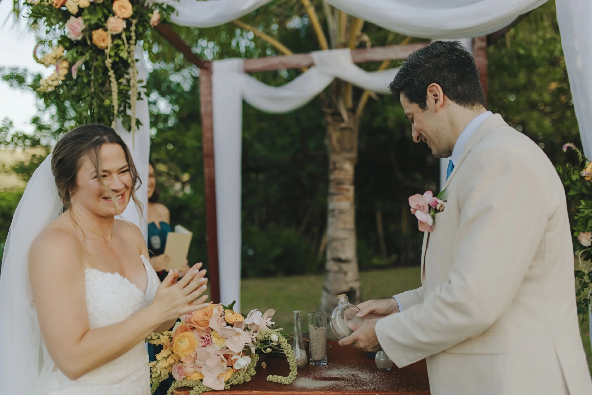 Bride laughing during a sand ceremony at her Punta Cana garden wedding as the wind catches her hair, groom pouring sand into a glass vessel