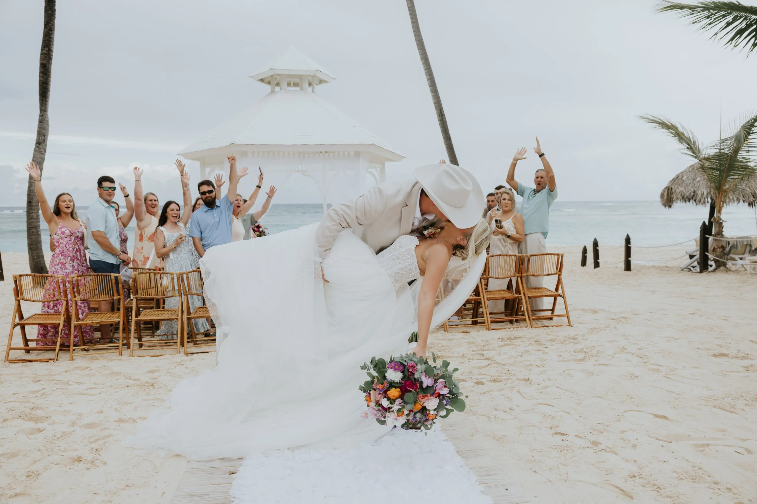 Groom dips bride for a kiss as cheering guests raise their arms along the beach aisle after the ceremony at Majestic Elegance Punta Cana