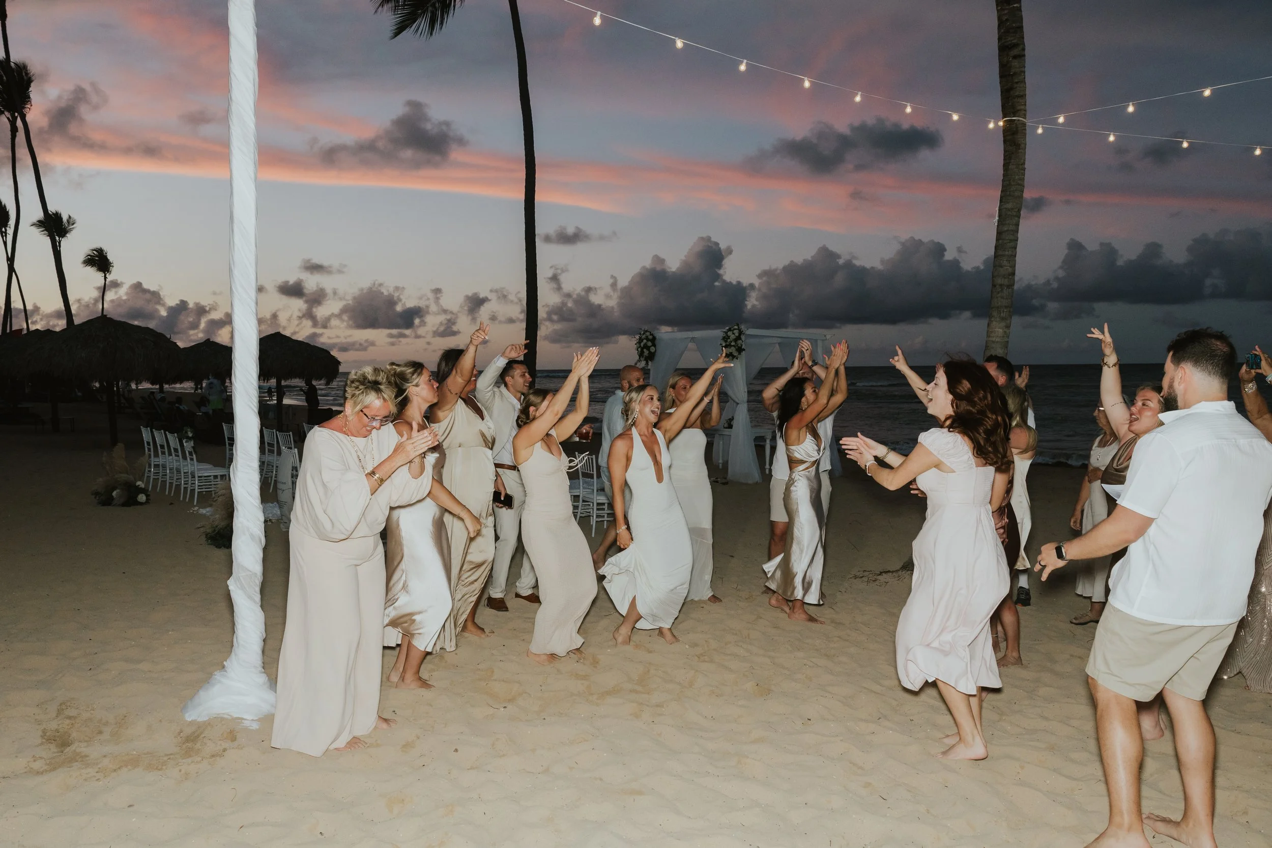 Wedding guests dancing with arms raised on the sand under string lights and a pink and purple sunset sky during a beach reception at Secrets Tides Punta Cana