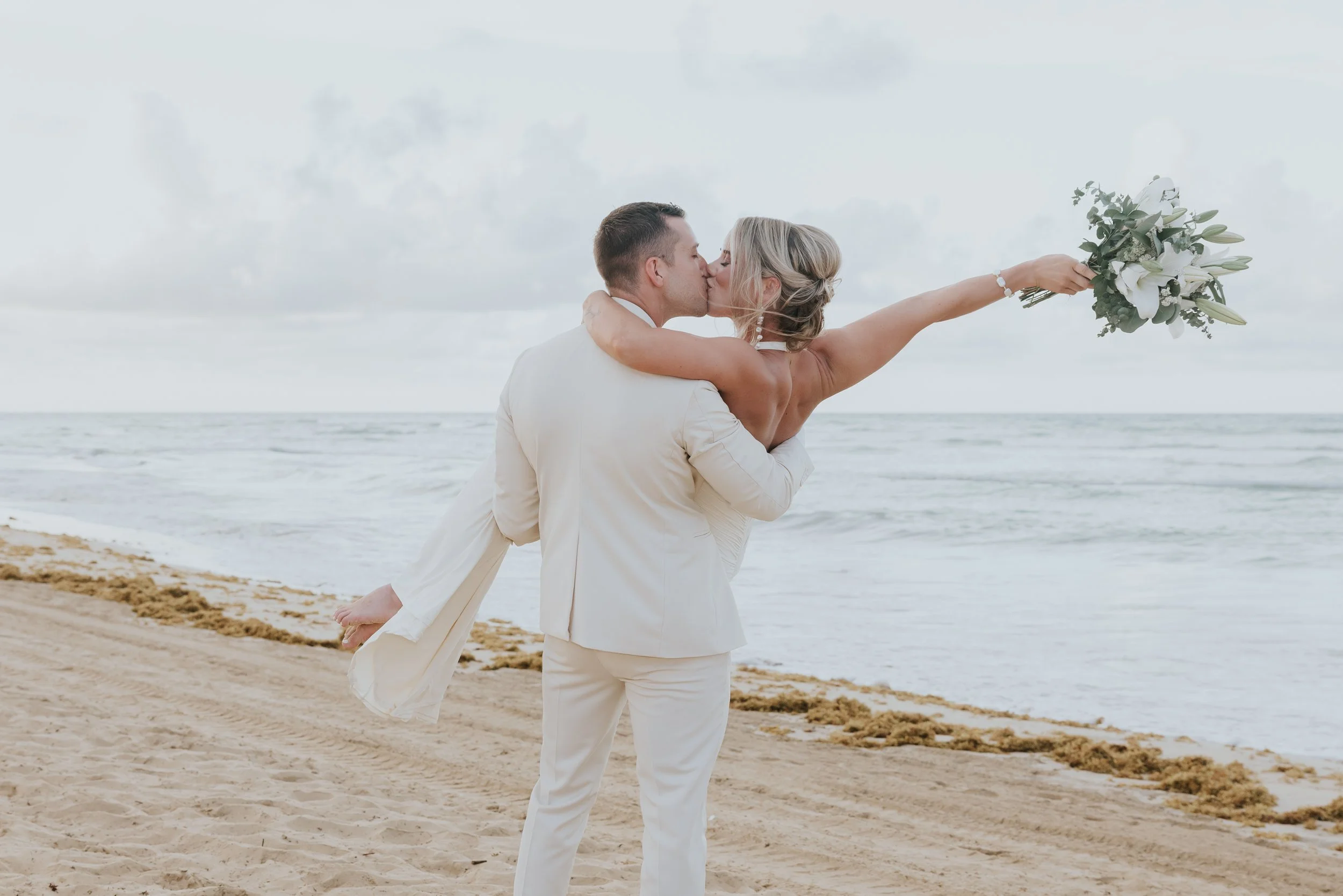 Groom lifting his bride in a kiss on the Secrets Tides shoreline, her arm stretched out holding a white and green bouquet, with the ocean behind them