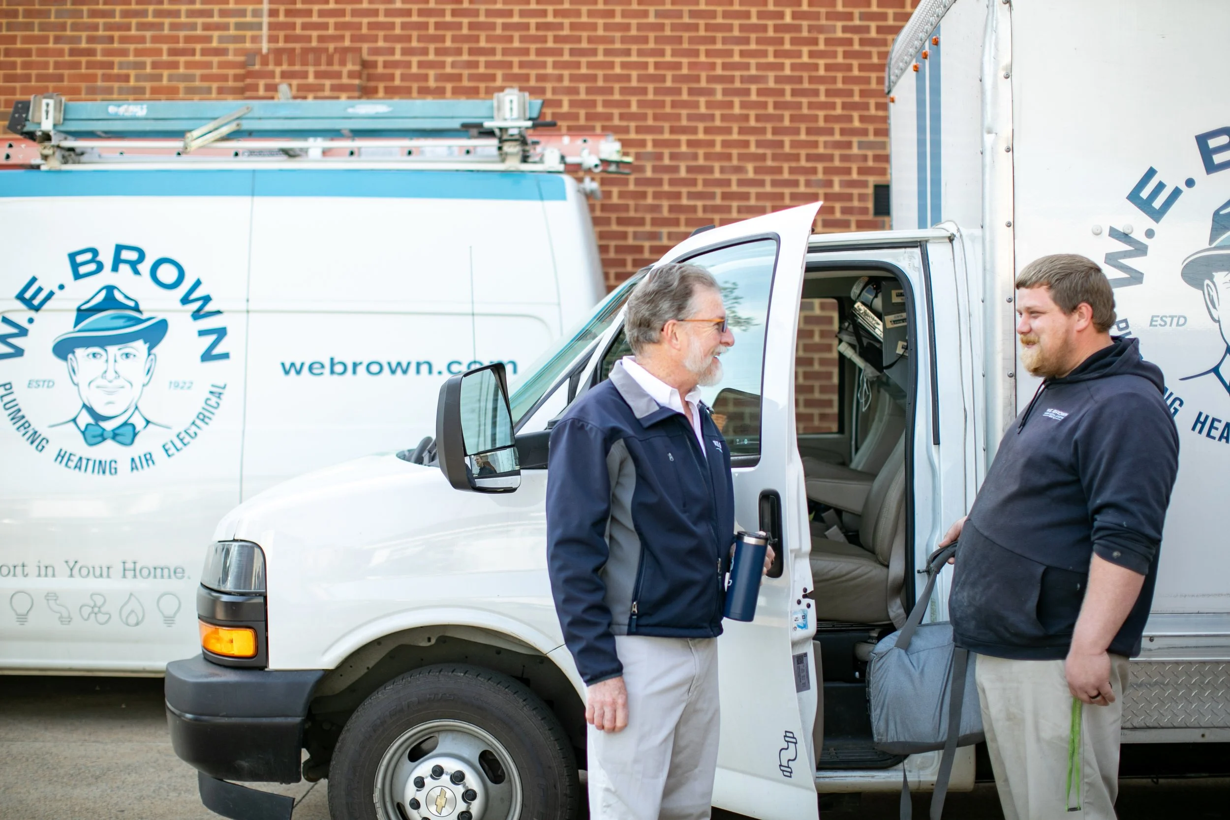 Two men standing near a white company van, smiling and talking. The van has a logo with a man's face in a hat, and the website 'webrown.com'. One man is holding a tumbler, and the other has a backpack.