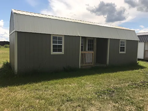 Wooden cabin with windows, a porch, and a metal roof in a grassy area under a cloudy sky.