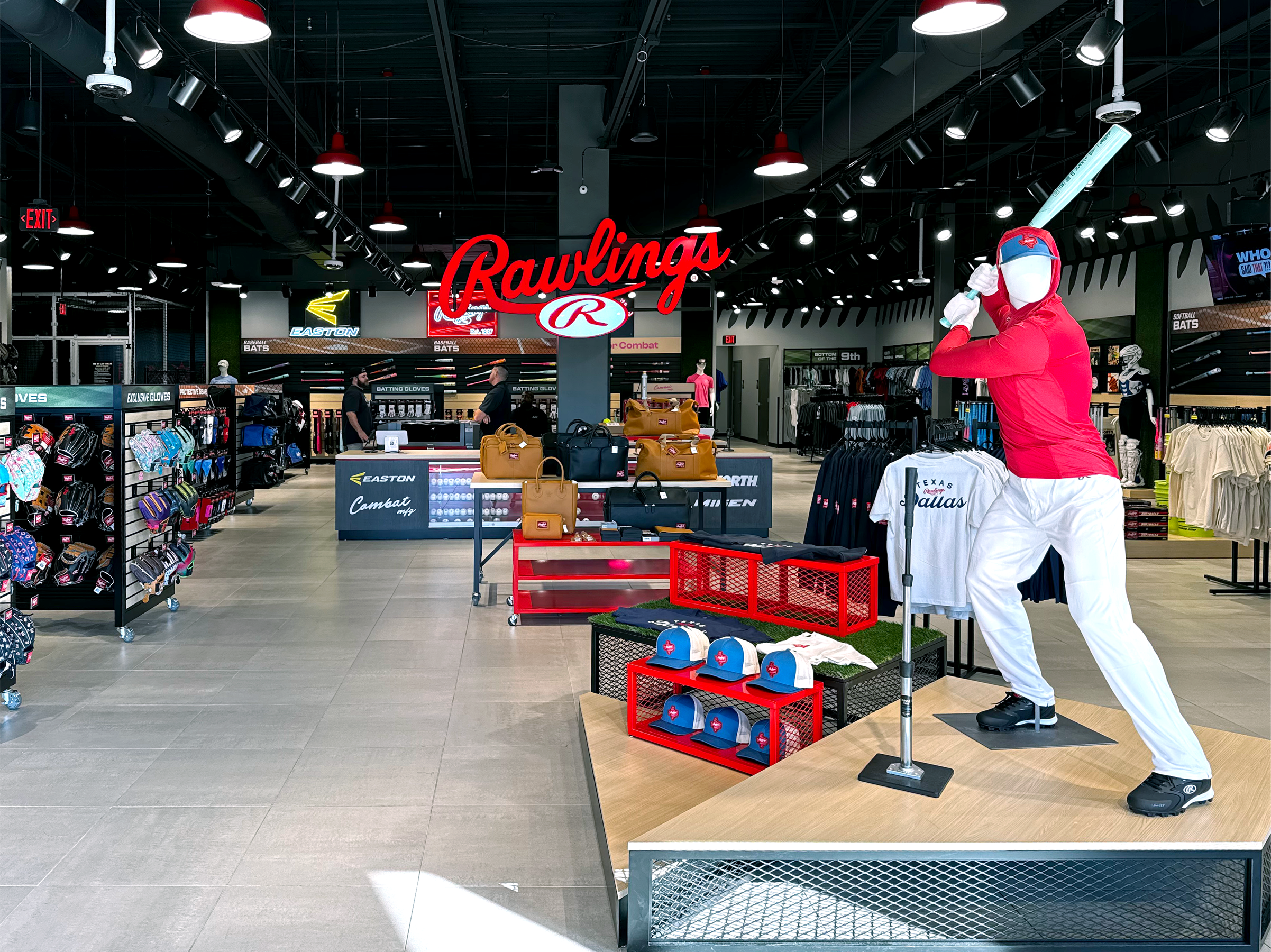 Inside a sporting goods store with a large red and white 'Rawlings' sign. There are various baseball gloves, bags, and apparel displayed. A mannequin dressed as a baseball player in a red shirt and white pants, holding a bat, is positioned on a platform.