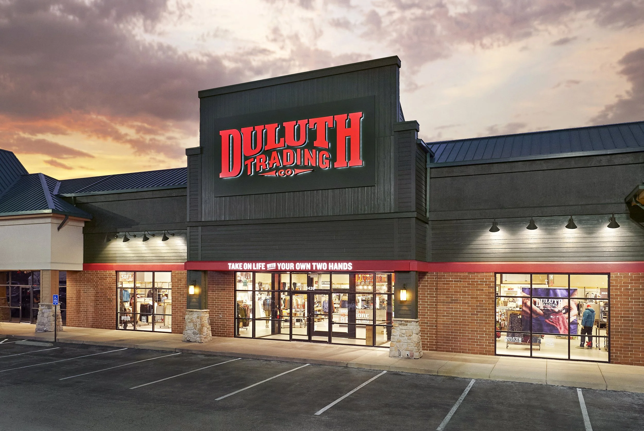 Exterior of Duluth Trading Co. store at sunset, with illuminated sign and parking lot in front.