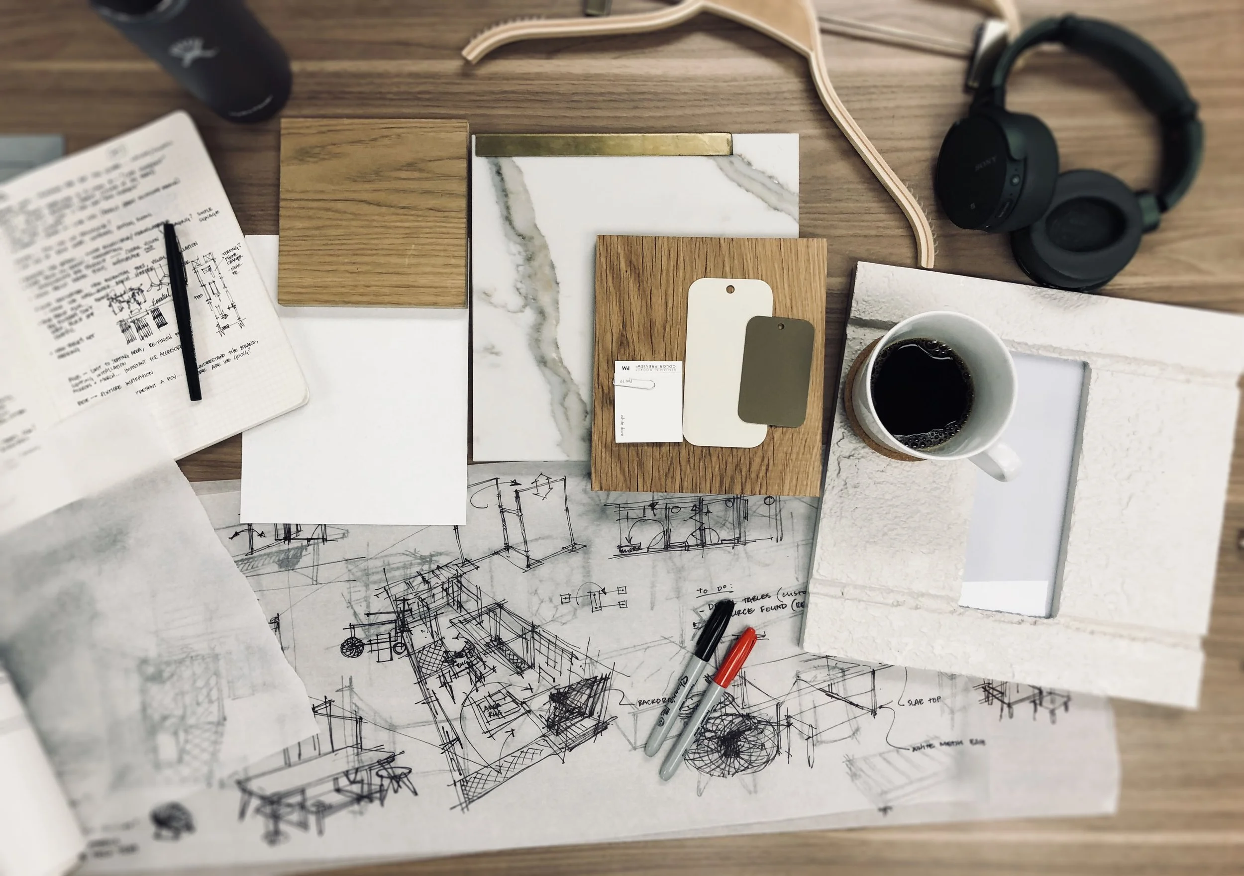 A wooden table with design sketches, color samples, a coffee cup, a notebook, a pair of black headphones, and various material samples for interior design.