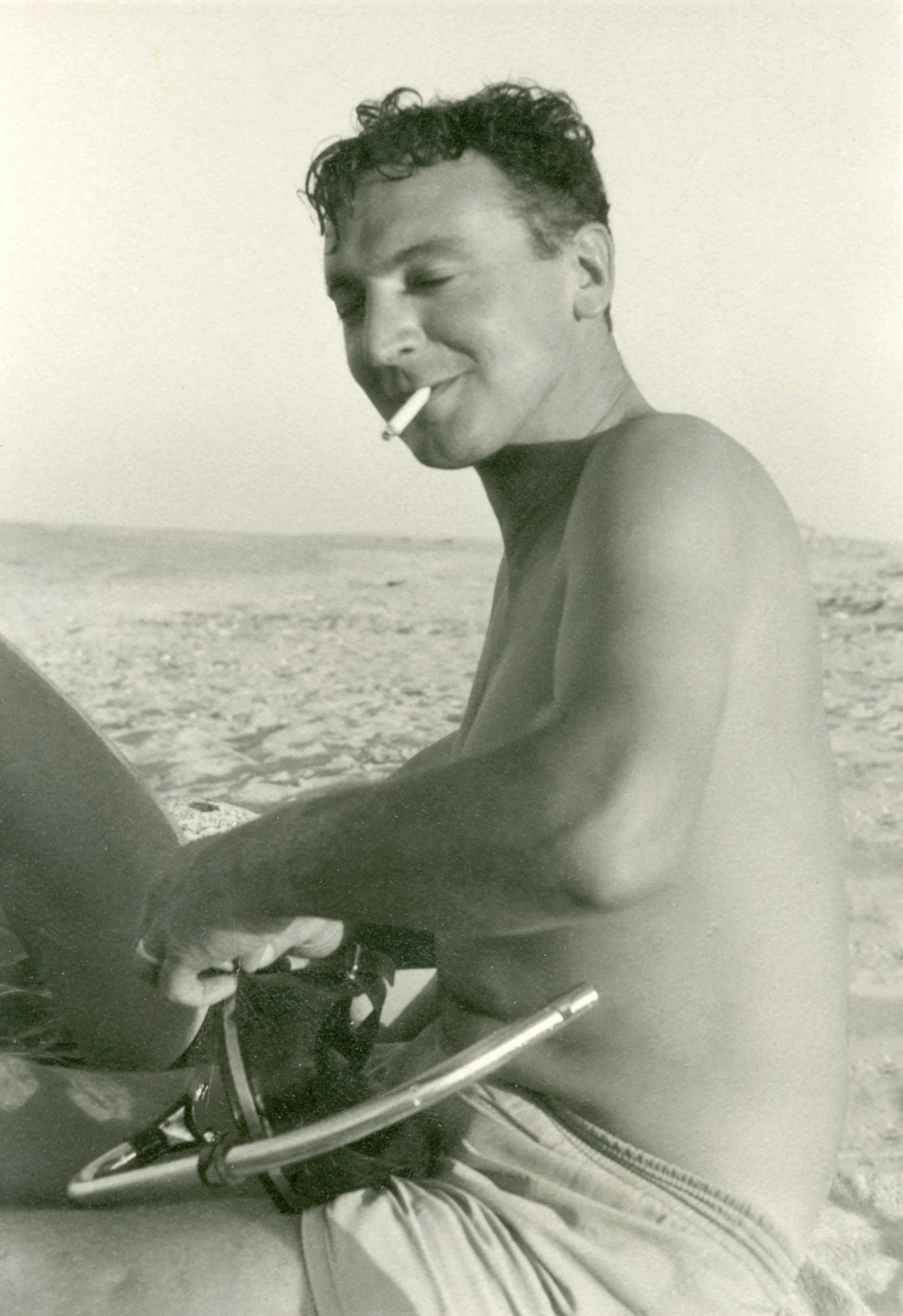 A black-and-white photo of a man with curly hair, shirtless, smoking a cigarette, sitting at the beach with a sandy shoreline in the background.