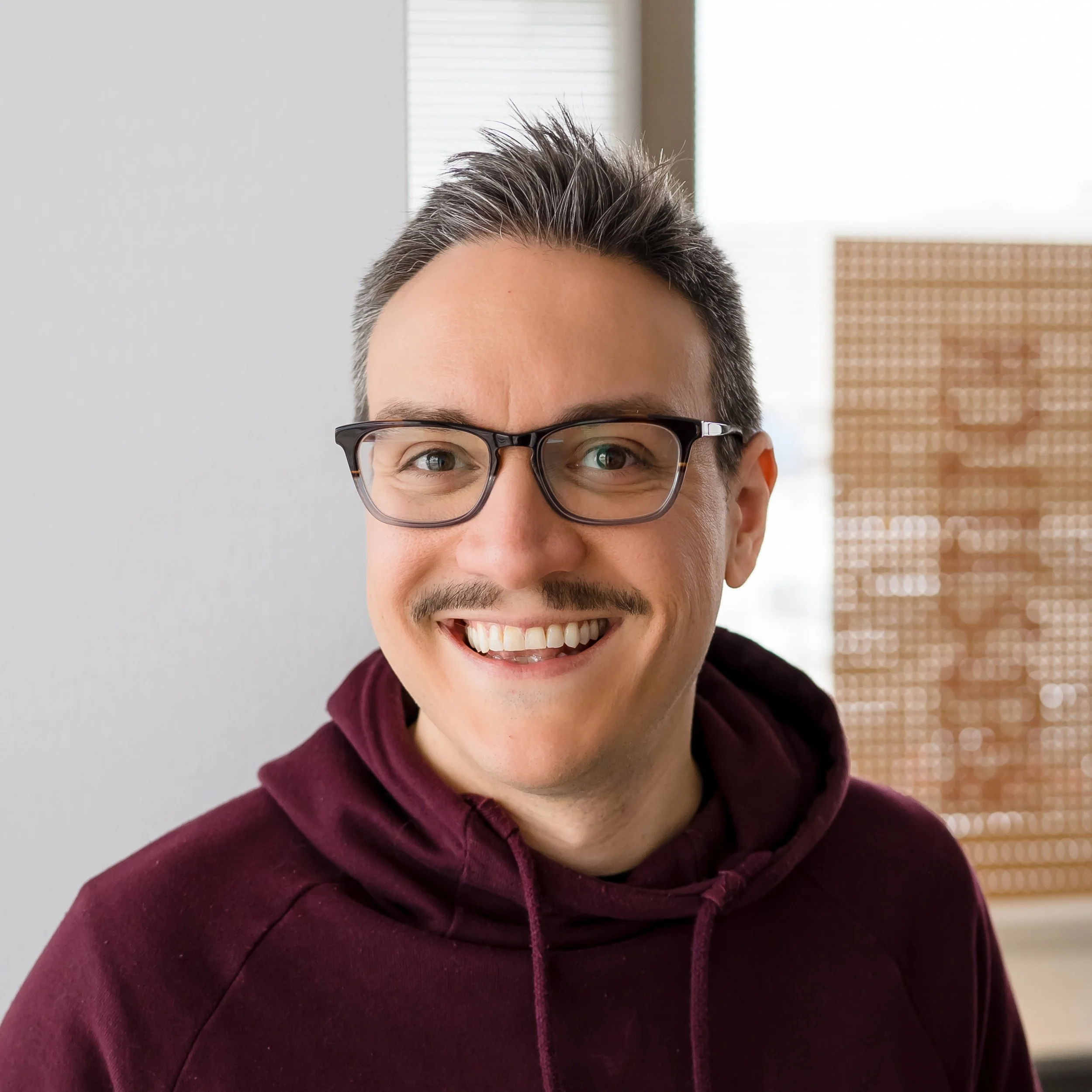 Young man wearing black shirt and black cap working on digital drawing tablet at a desk in front of large window with blinds during daylight.
