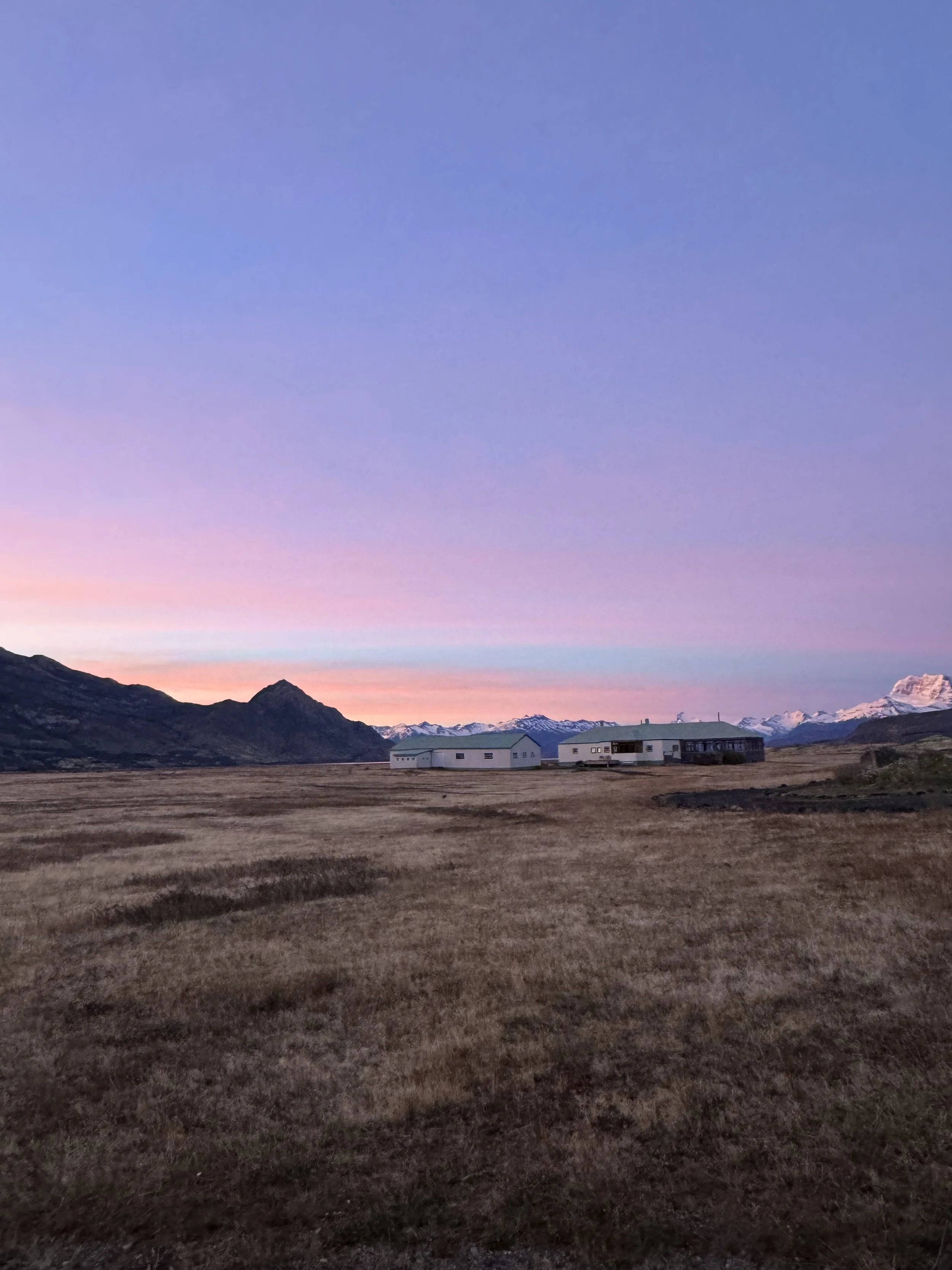 A rural landscape with mountains in the background, a small white building, and a clear sky with pink and purple hues at sunset.