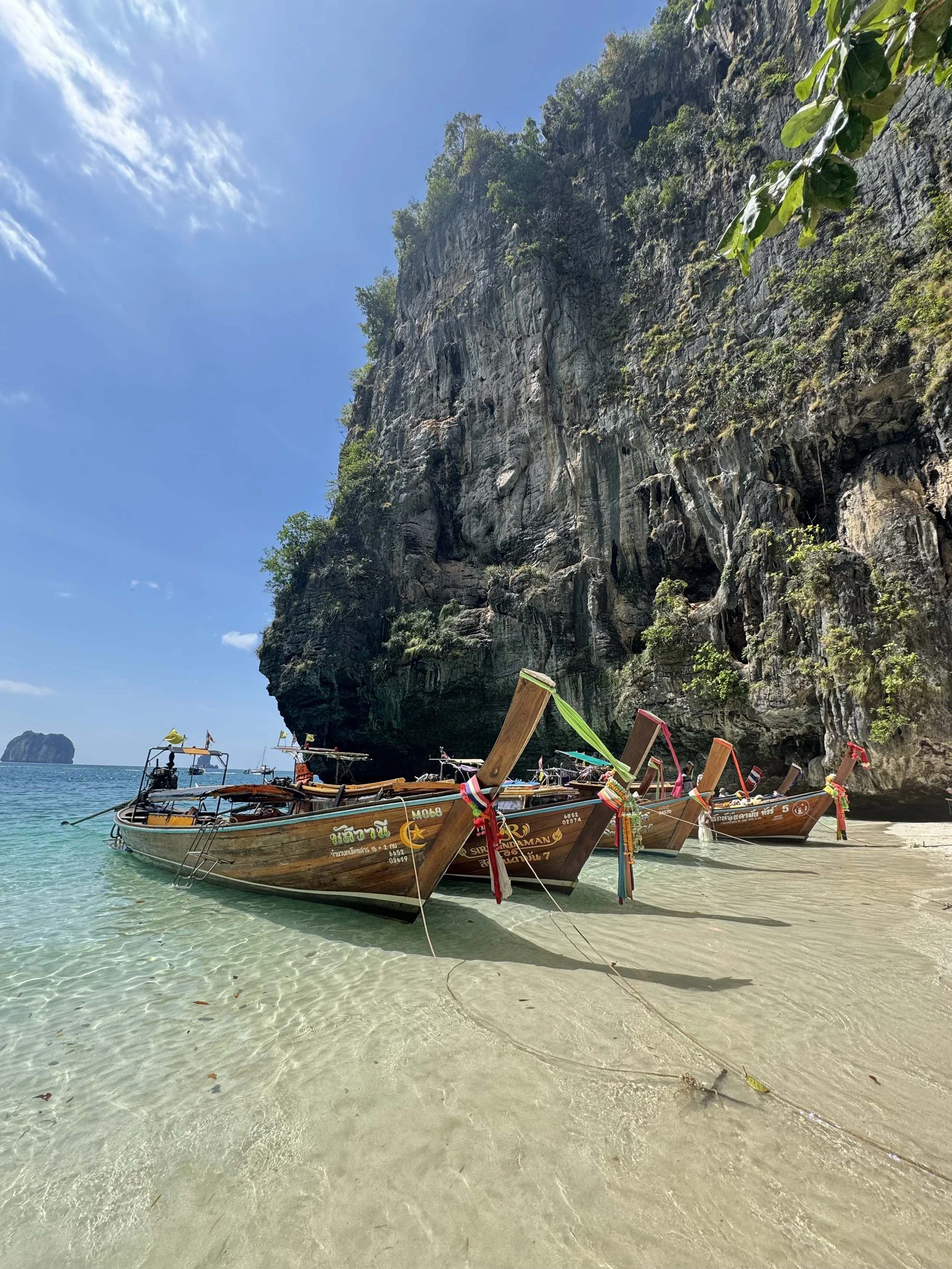 Boats on a sandy beach at the base of a large rocky cliff with greenery, under a clear blue sky.