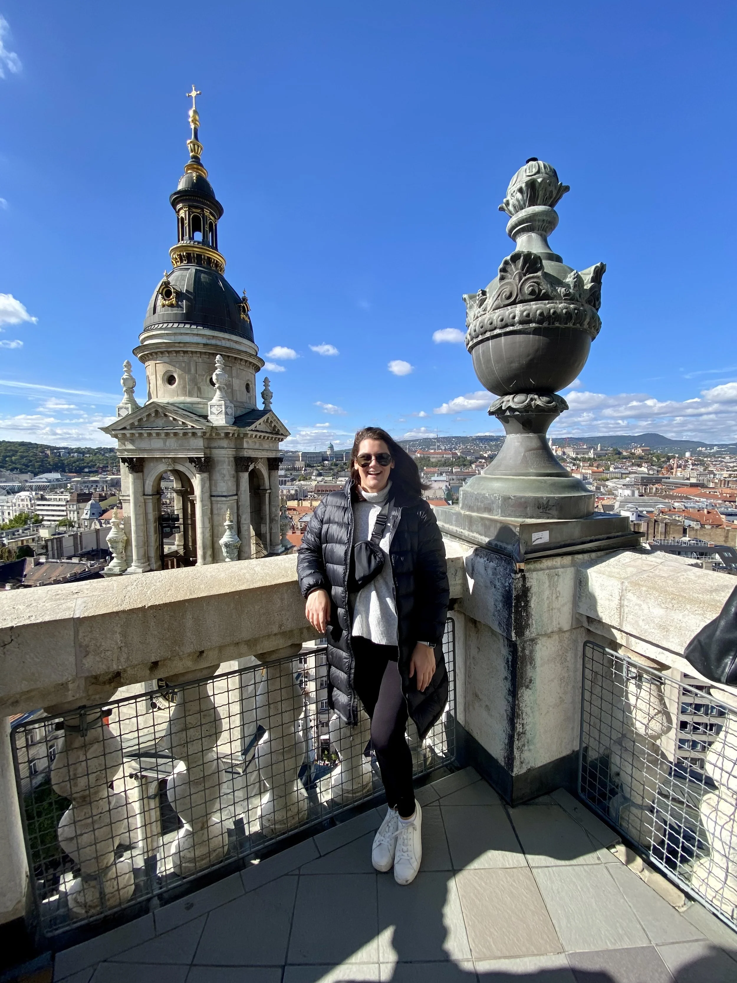 A woman smiling on a rooftop with a view of a city skyline, church steeple, and decorative urns, wearing sunglasses, a puffy jacket, gray sweater, black pants, and white sneakers.