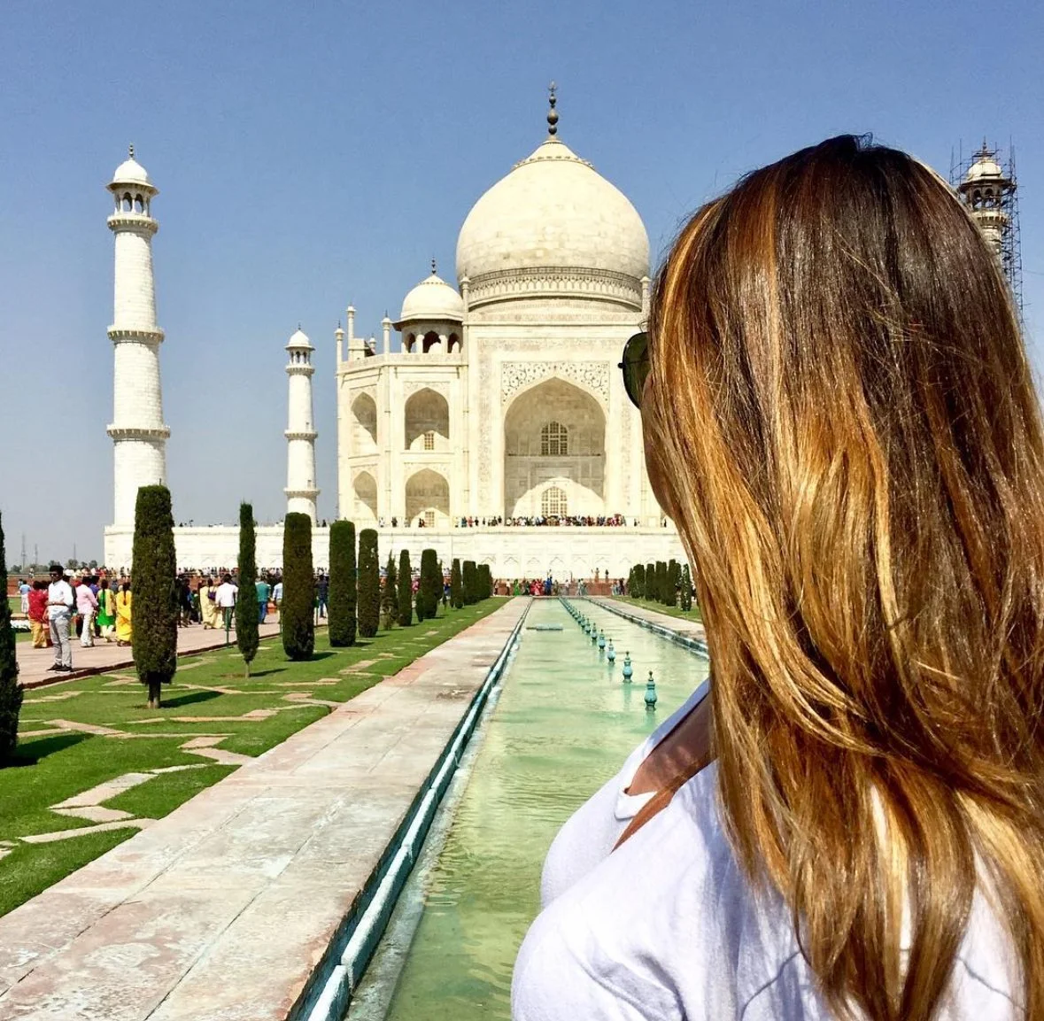 A woman with brown hair and sunglasses standing near a water channel, with the Taj Mahal in the background on a clear day.