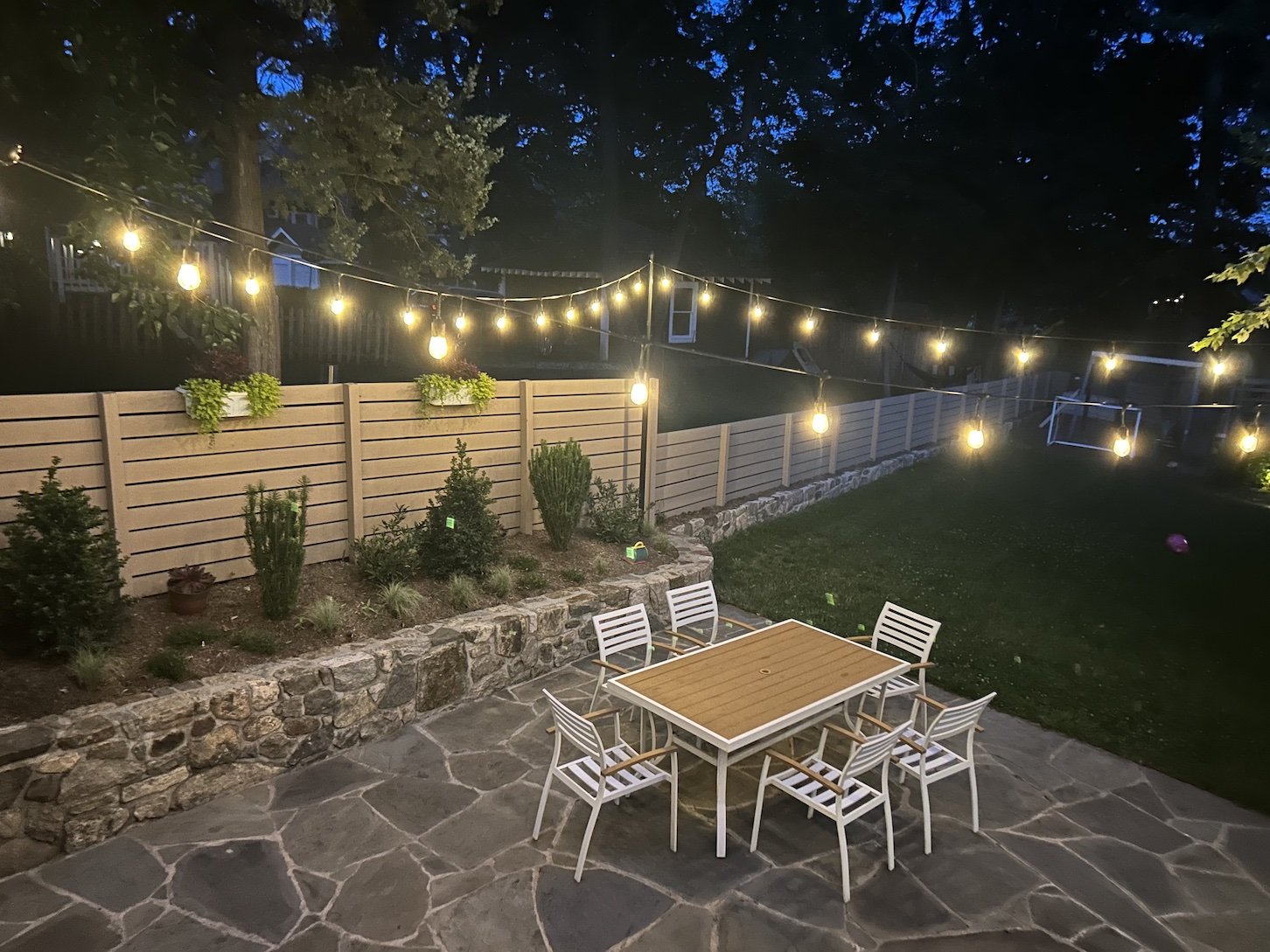 Back patio lighting with italian string lights above neutral colored patio furniture, a natural stone patio and a wooden fence.jpg
