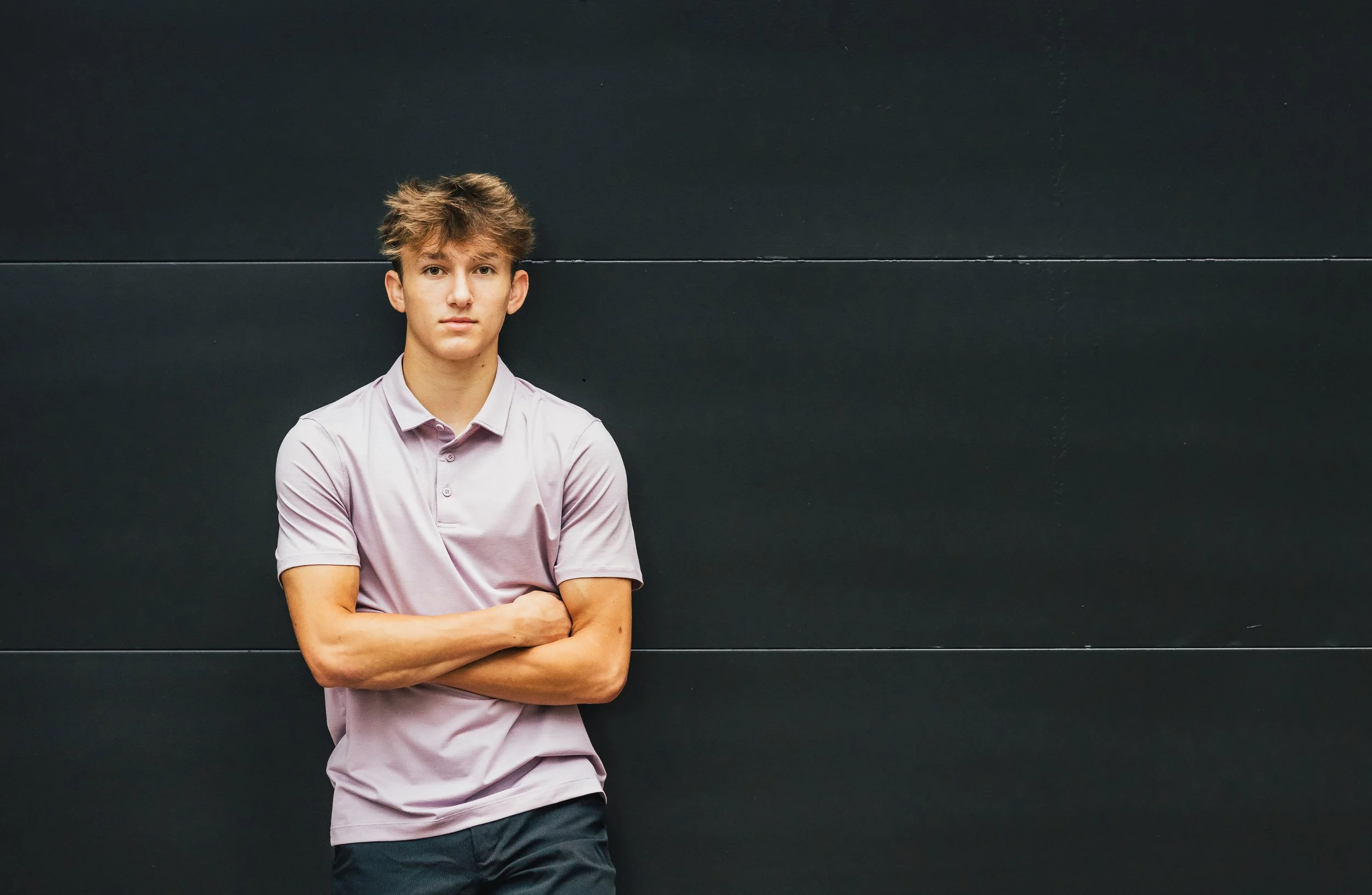 Young man with short, tousled hair wearing a light pink polo shirt and dark shorts, standing against a dark wall with his arms crossed.