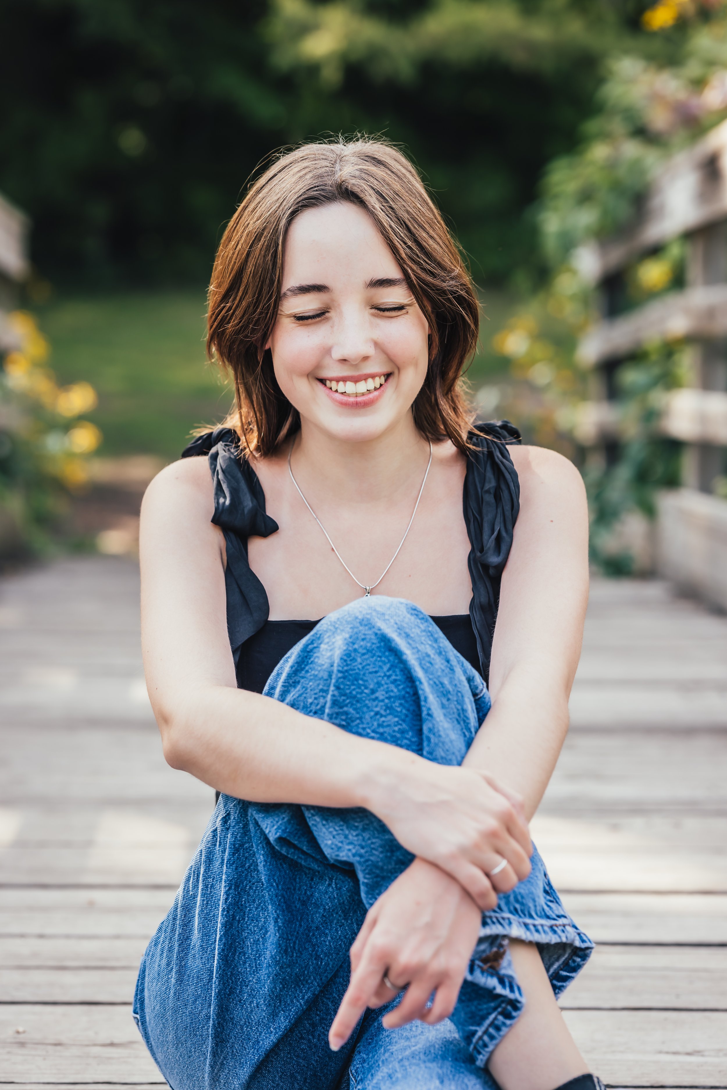 A young woman with brown hair smiling sitting on a wooden dock in a park or garden during daytime