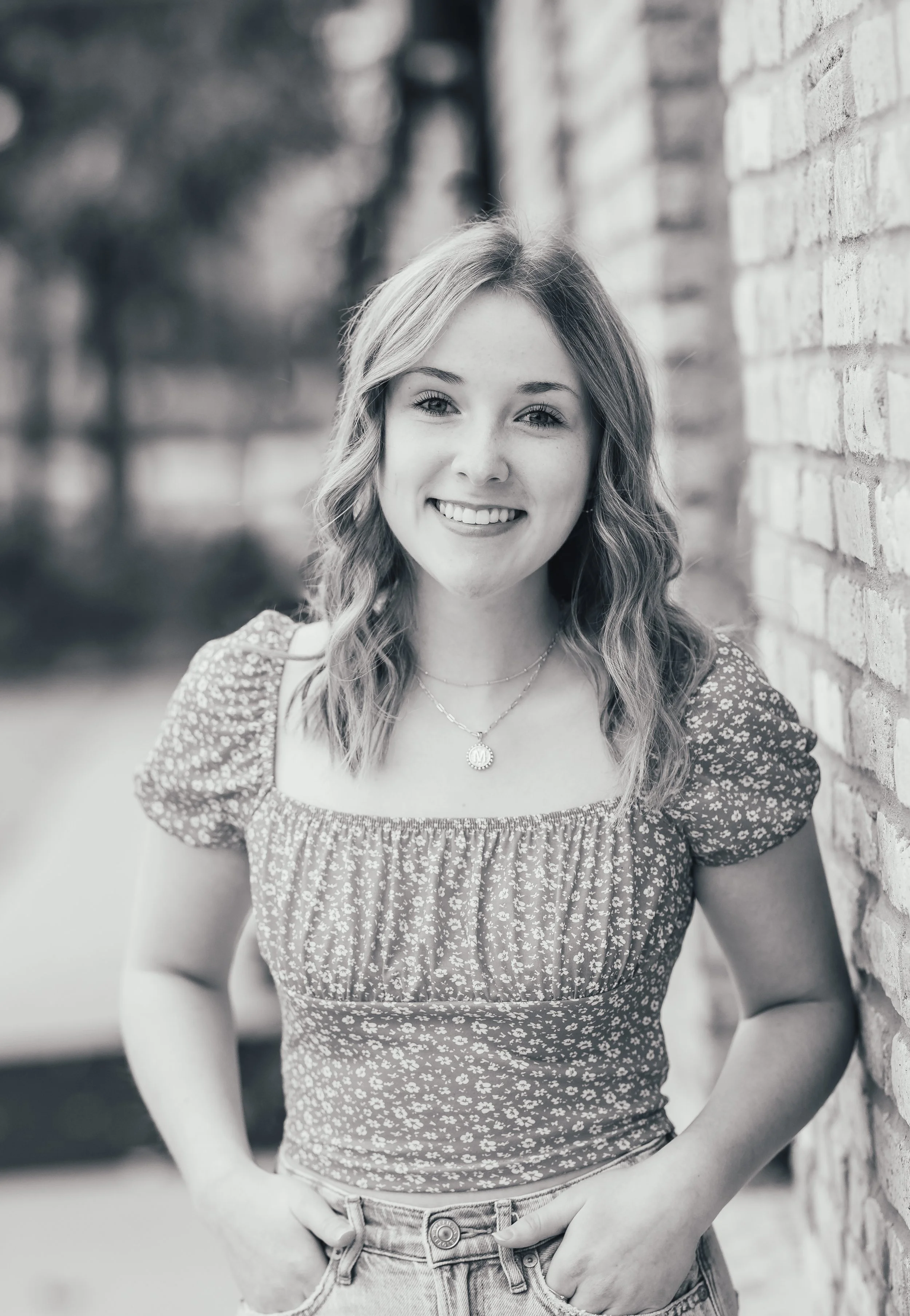 A young woman with wavy hair smiling and standing next to a brick wall outdoors.