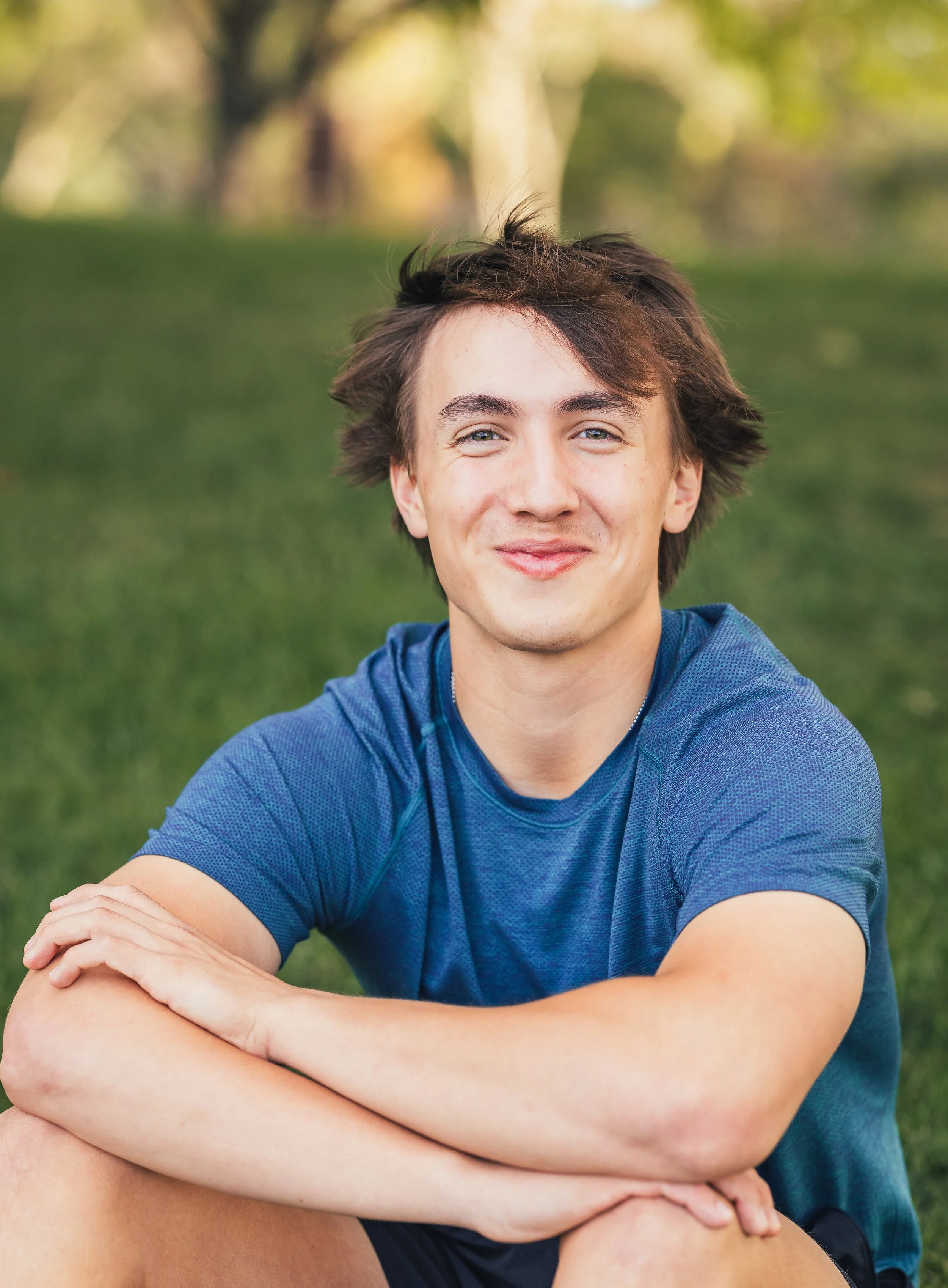 A young man with brown hair, blue eyes, wearing a blue athletic shirt, sitting outdoors on grass with trees in the background, smiling at the camera.