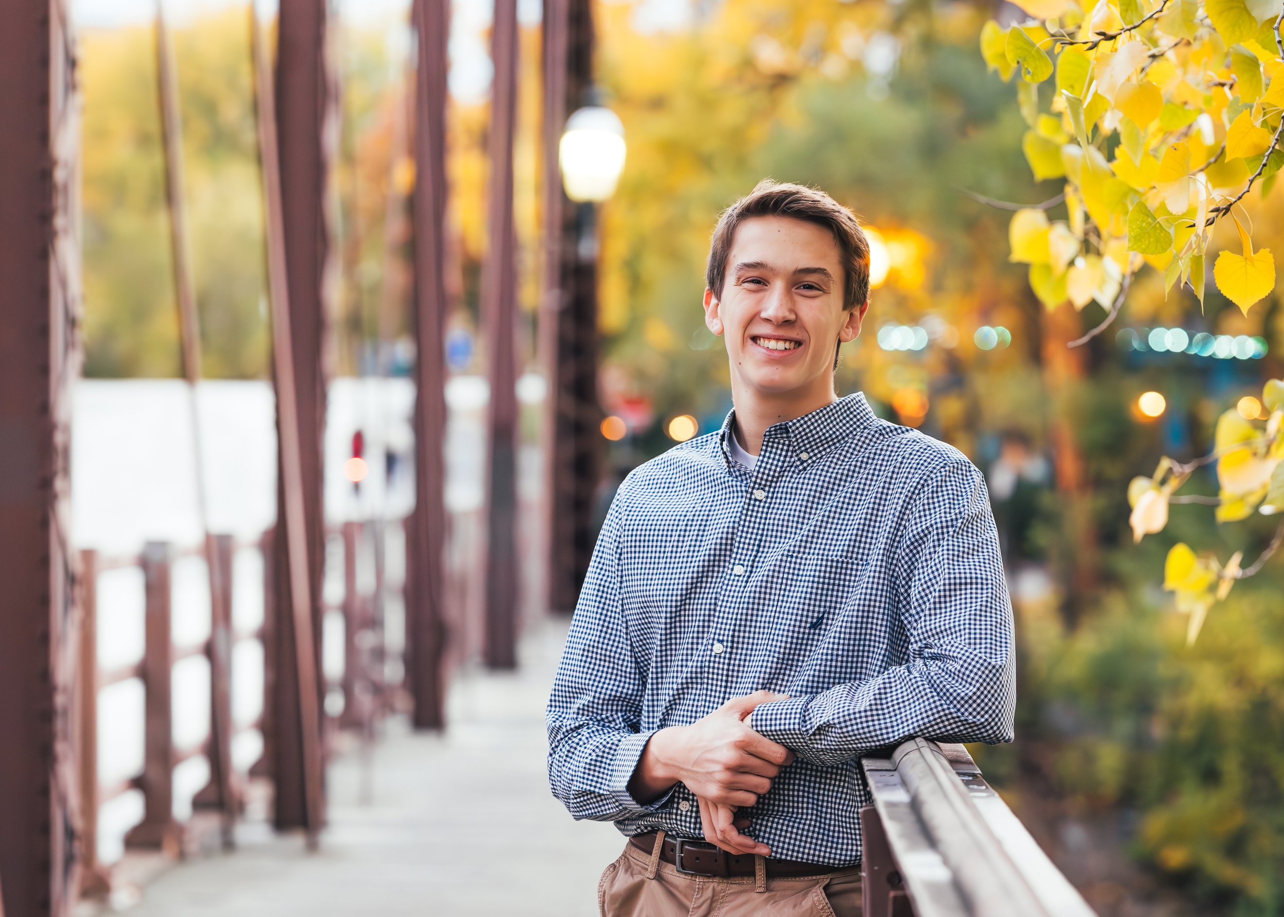 A young man with brown hair and a checkered shirt smiling outdoors on a bridge with autumn trees in the background.