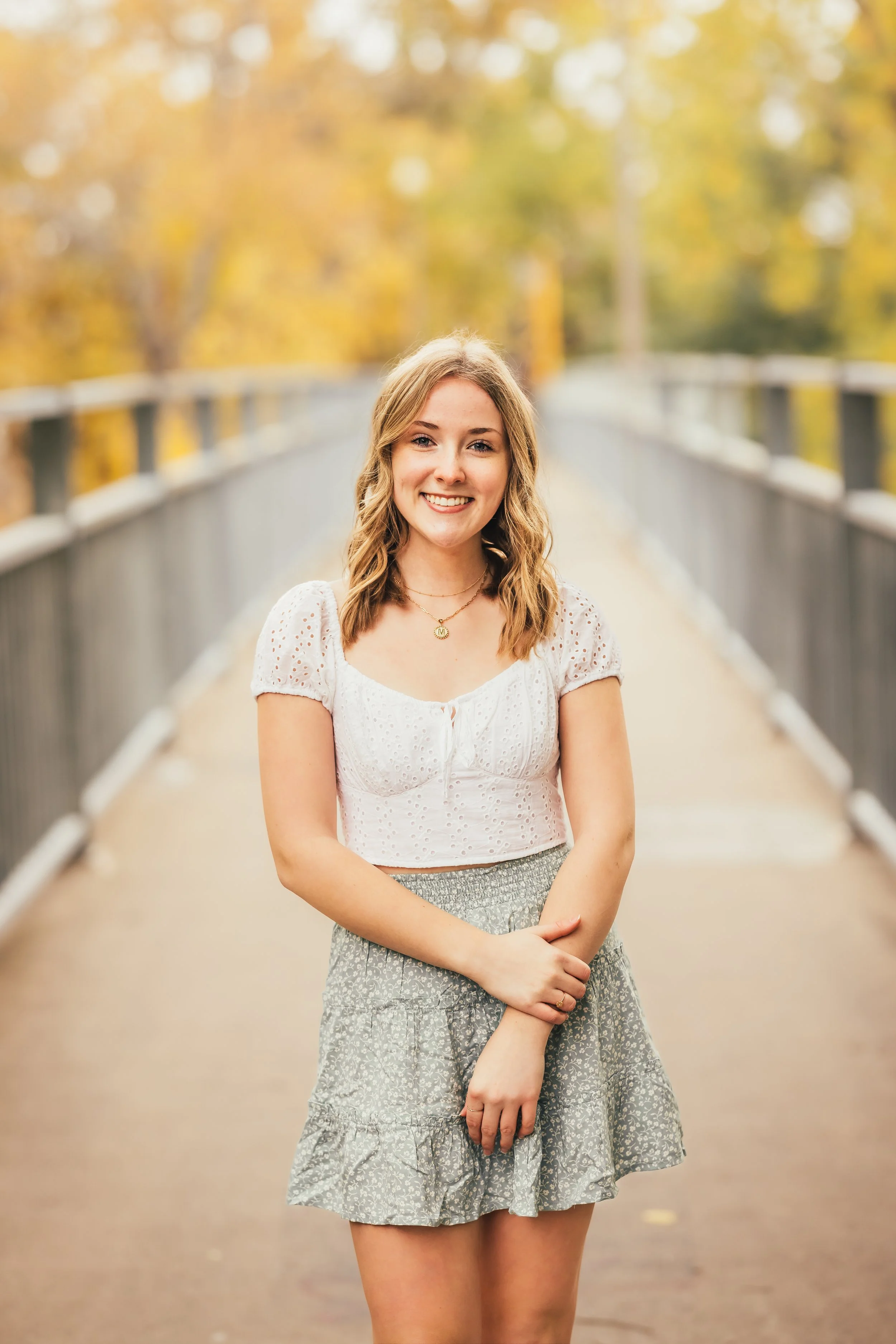 A young woman in a white eyelet crop top and a gray floral skirt standing on a bridge during fall, smiling at the camera with autumn trees in the background.