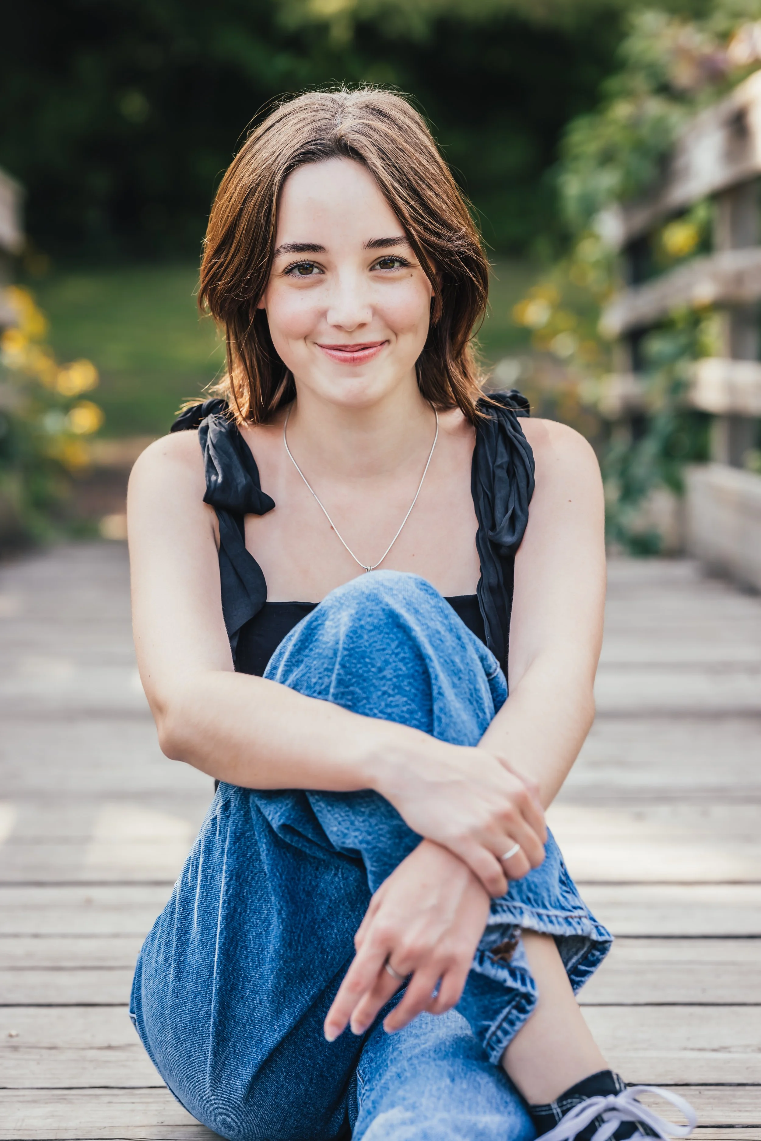 A young woman with short brown hair sitting outdoors on a wooden dock, smiling at the camera, dressed in a black sleeveless top and blue jeans.