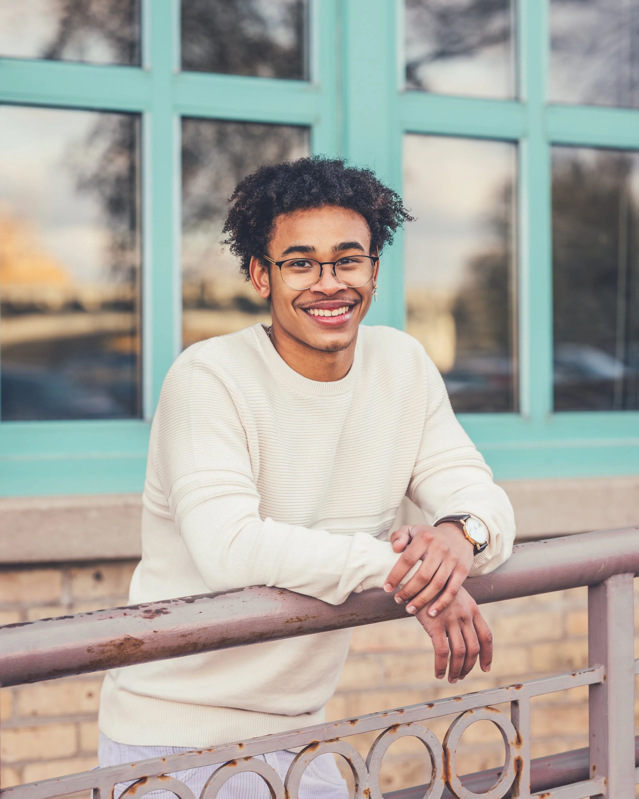 Smiling young man with curly hair, glasses, and earrings leaning on a metal railing outside a building with large teal-framed windows.