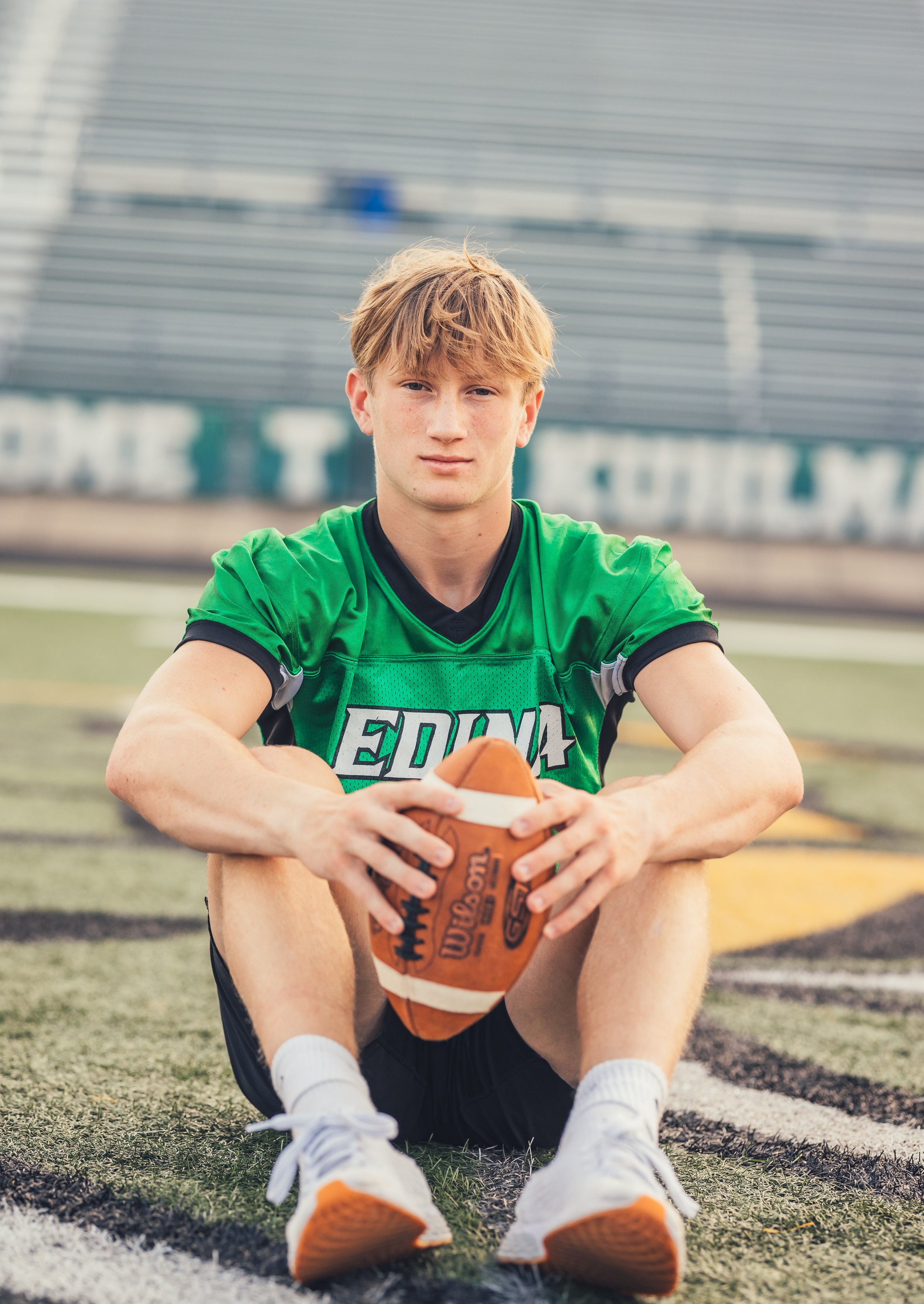 A teenage boy in a green football jersey sitting on a football field, holding a football, looking at the camera.