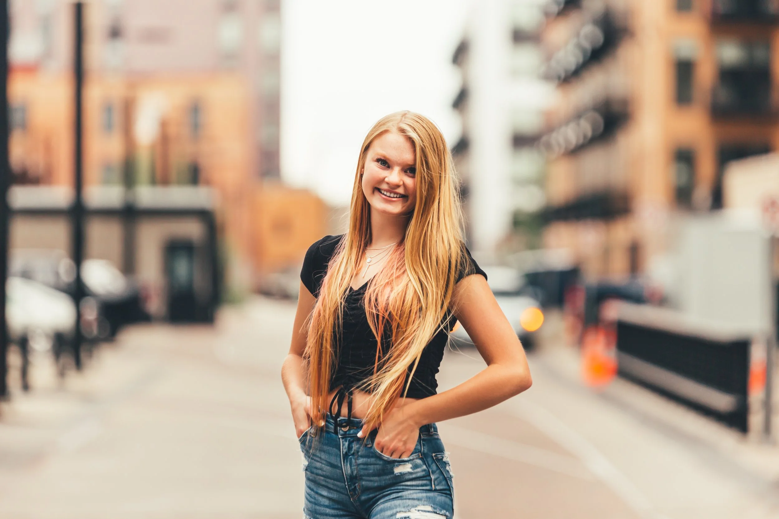 A smiling young woman with long red hair standing on a city street with blurred buildings and cars in the background.