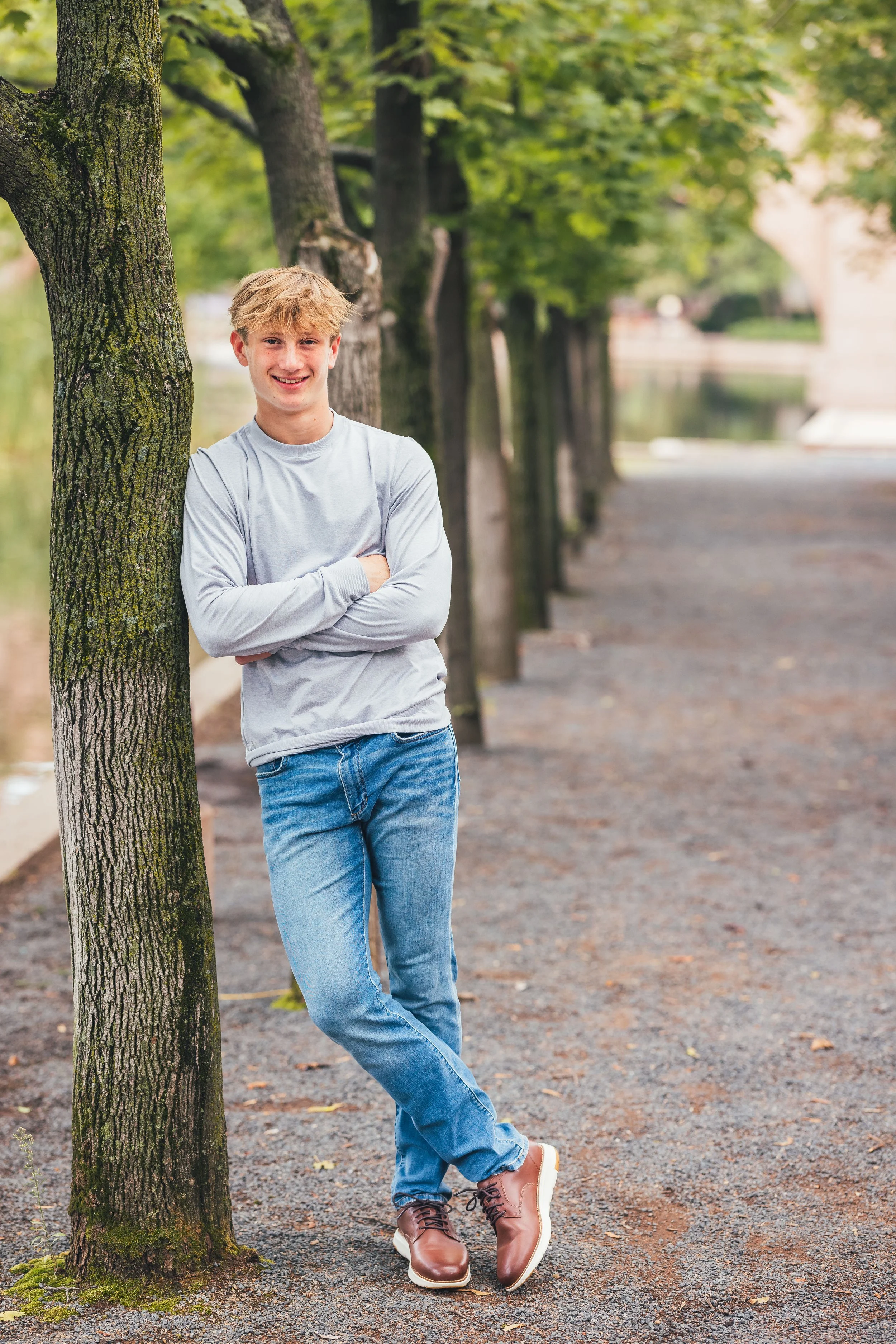 A young man with blond hair, wearing a gray long-sleeve shirt, blue jeans, and brown boots, leans against a tree on a pathway in a park with green trees and water in the background.