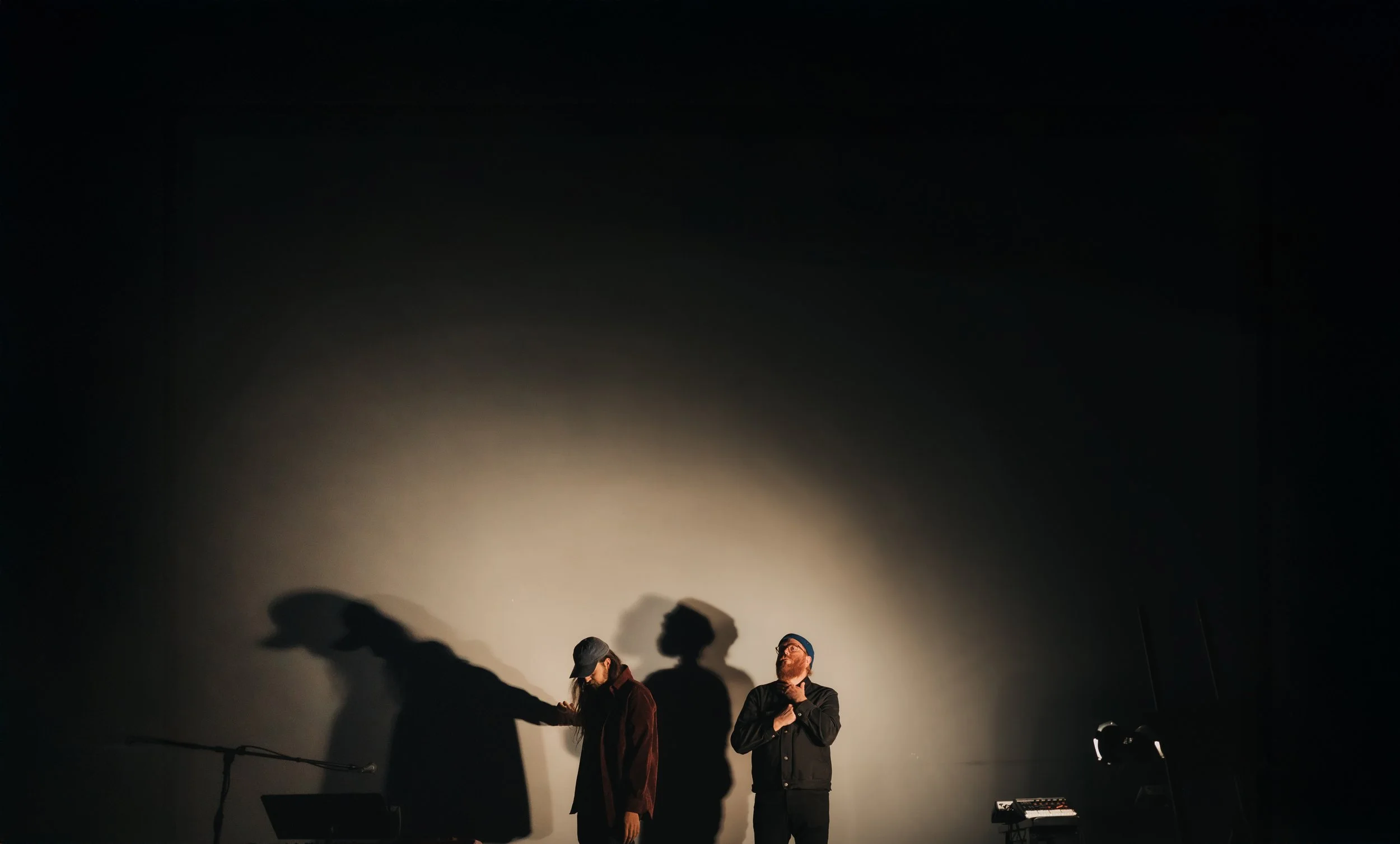 Two performers on a stage with their shadows cast on a blank wall behind them, with a microphone stand and electronic equipment nearby.