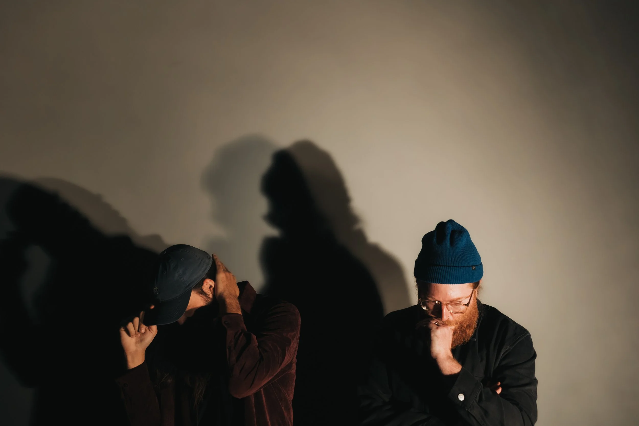 Three people with dark clothing and hats, standing in front of a plain wall, casting dramatic shadows behind them.