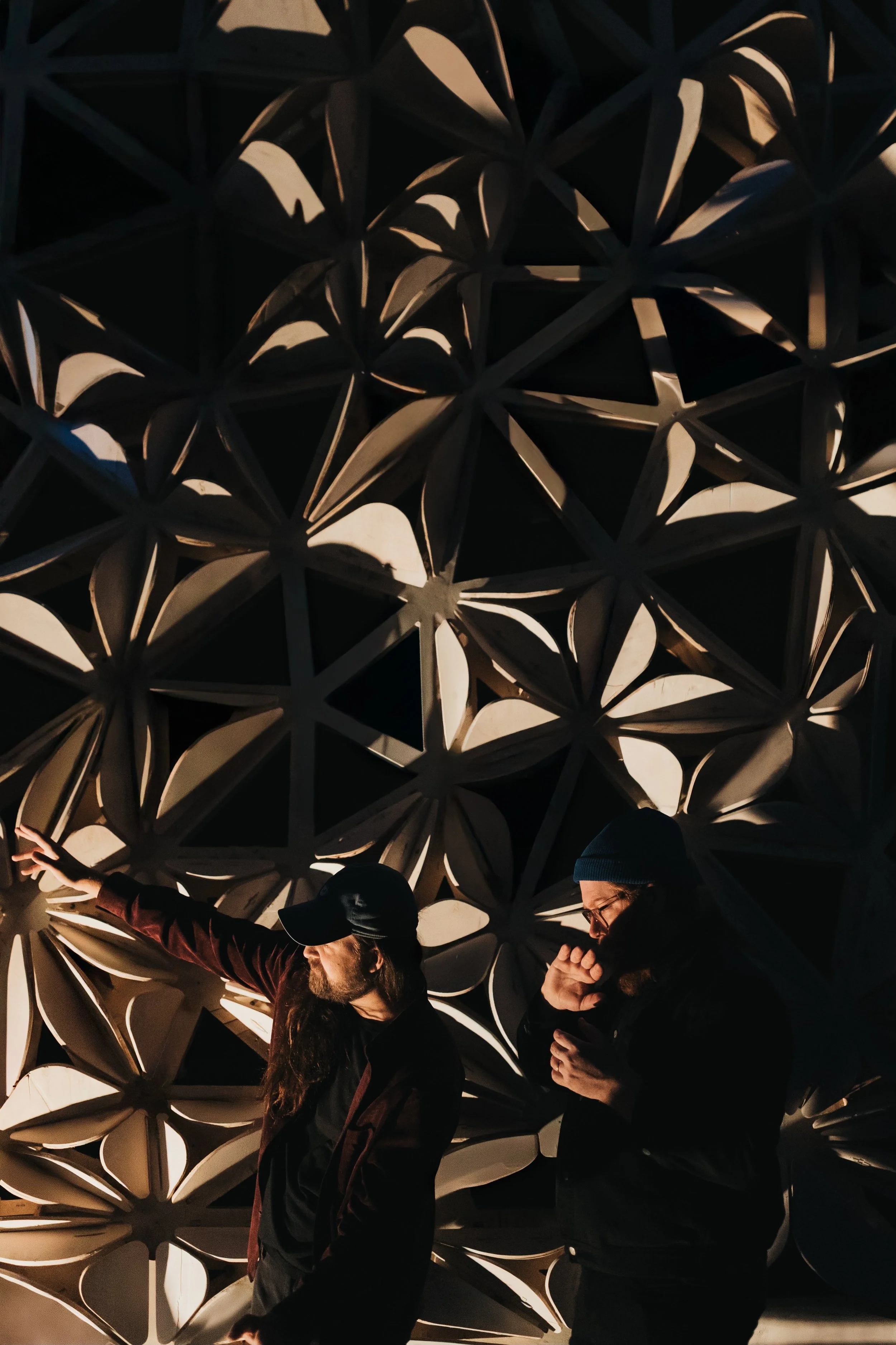 Two men standing in front of a large decorative lattice structure, one pointing towards it and the other looking at a phone, nighttime.