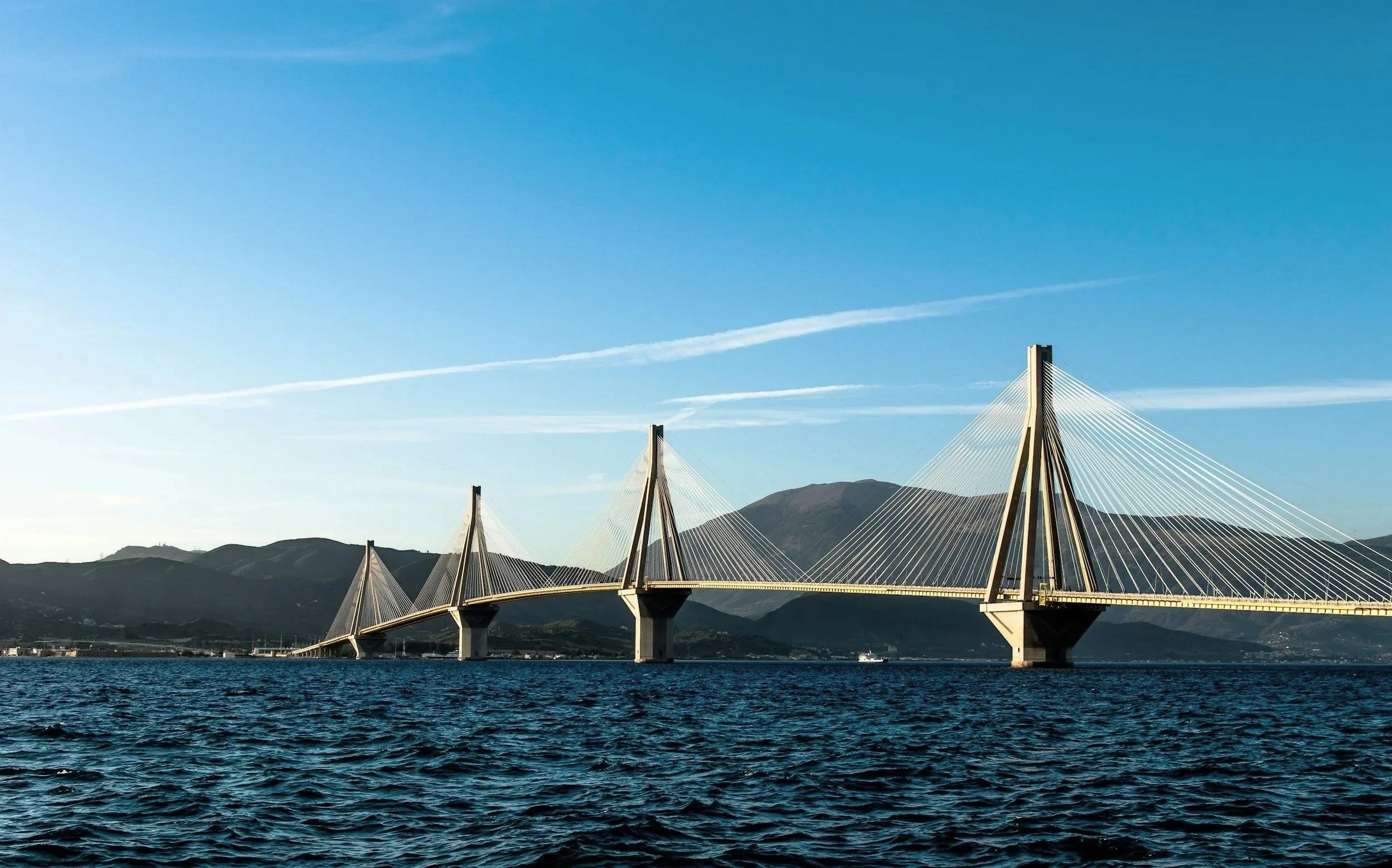 A modern cable-stayed bridge over a body of water with mountains in the background and a clear blue sky.