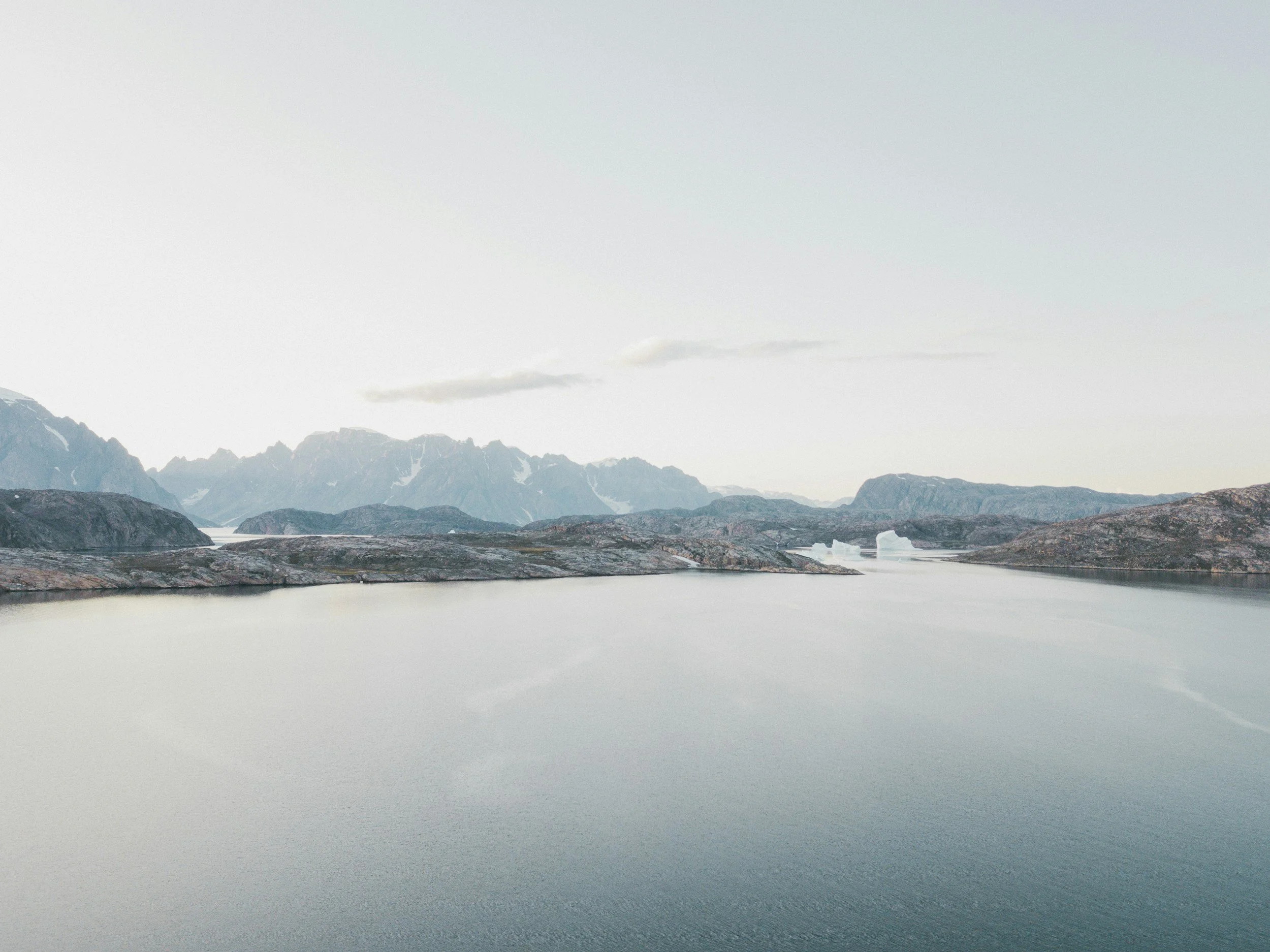 A scenic view of a calm lake with rocky islands, snow-capped mountains in the background, and a cloudy sky.