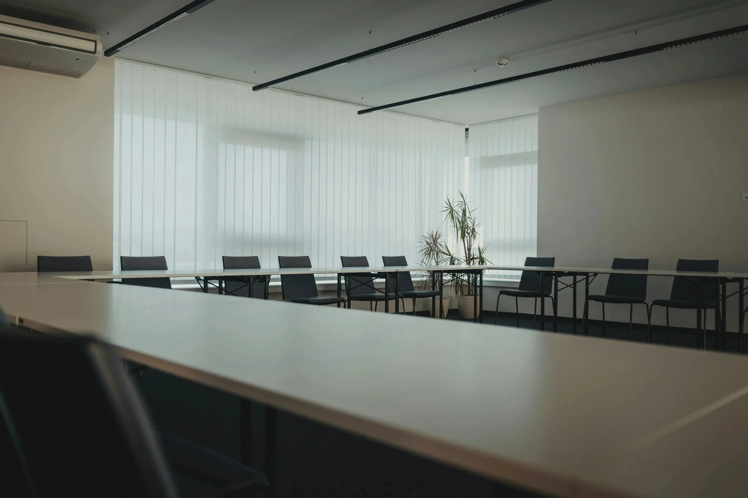 Empty conference room with white tables, black chairs, large windows with white blinds, potted plants, and ceiling lighting