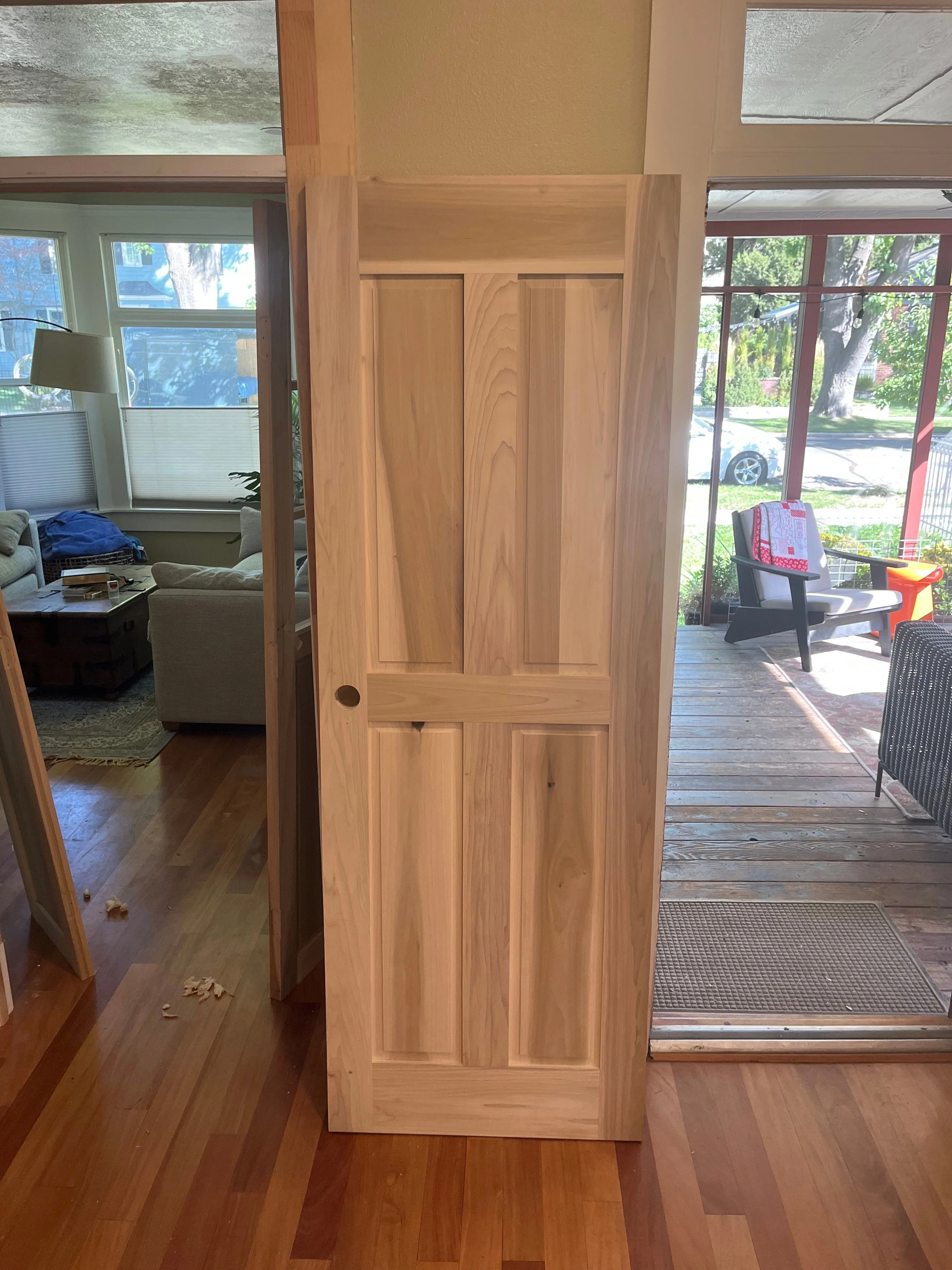 Unfinished wooden cabinet with four panels, standing in a room with hardwood floor, near a sliding glass door leading to a porch with chairs and a view of trees and a street.