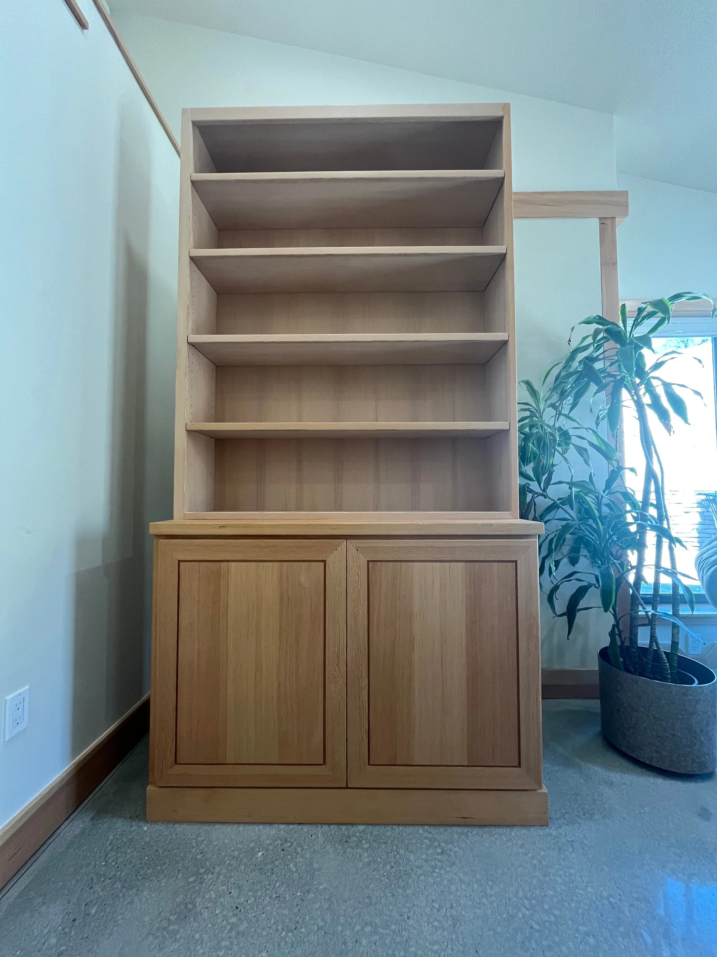 Unfinished wooden bookshelf with four shelves and cabinet doors at the bottom, placed in a room next to a potted plant near a window.