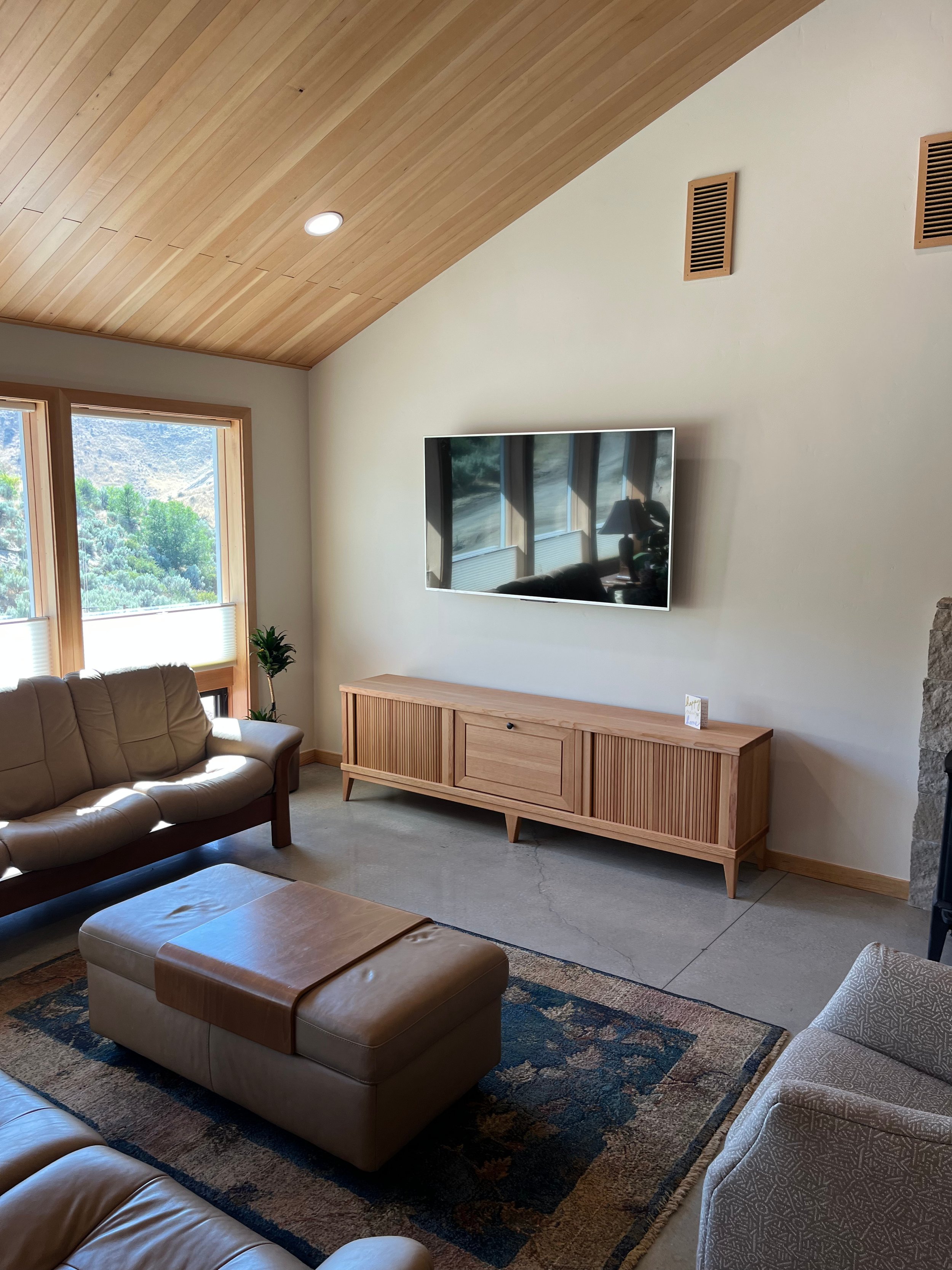Living room with beige leather couches, a wooden TV stand, a flat-screen TV, large windows showing greenery, a wooden ceiling, and area rugs.