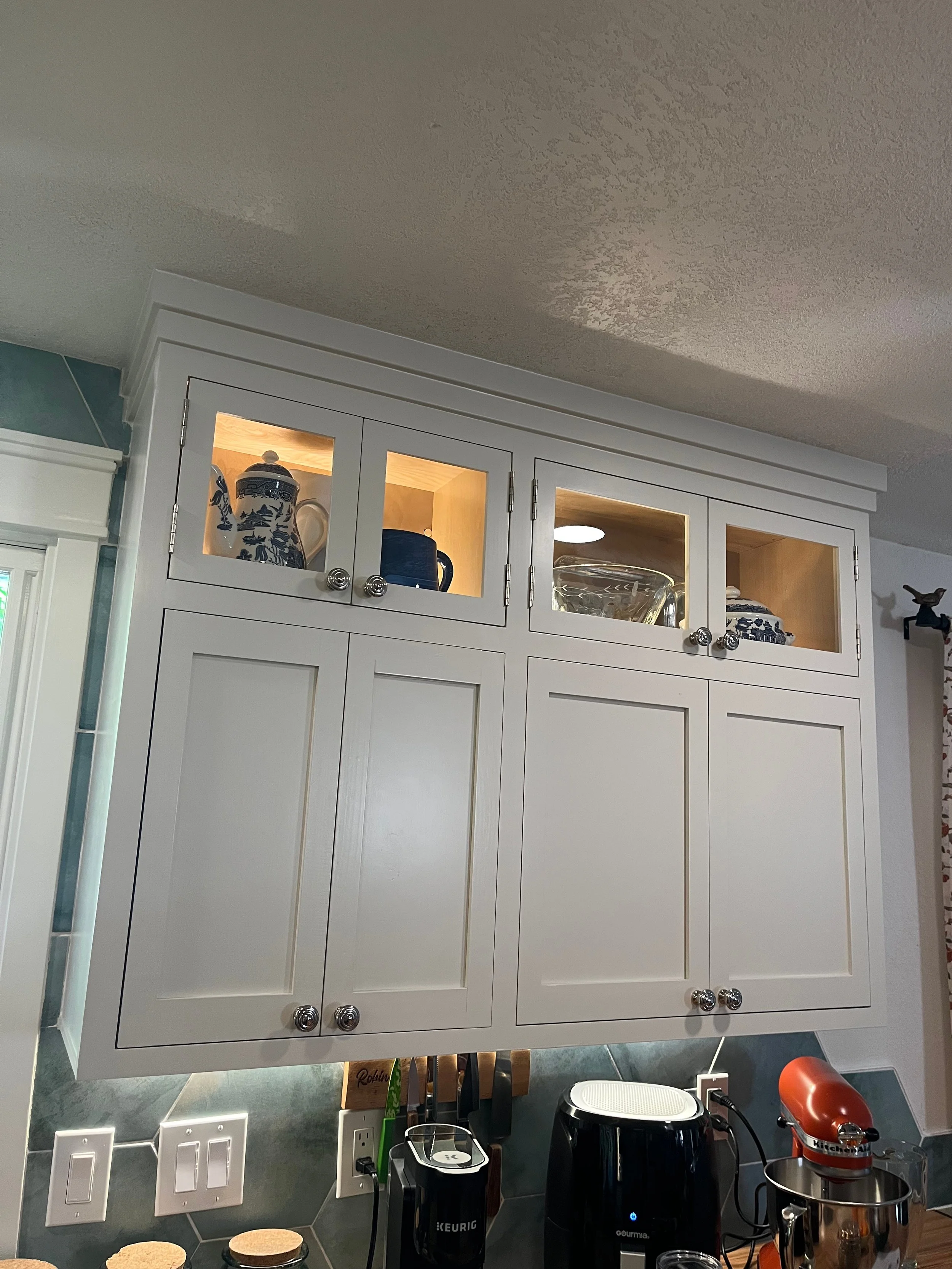 White kitchen cabinet with glass doors displaying blue and white dishes, above a dark countertop with small appliances, including a coffee maker, air fryer, and stand mixer.