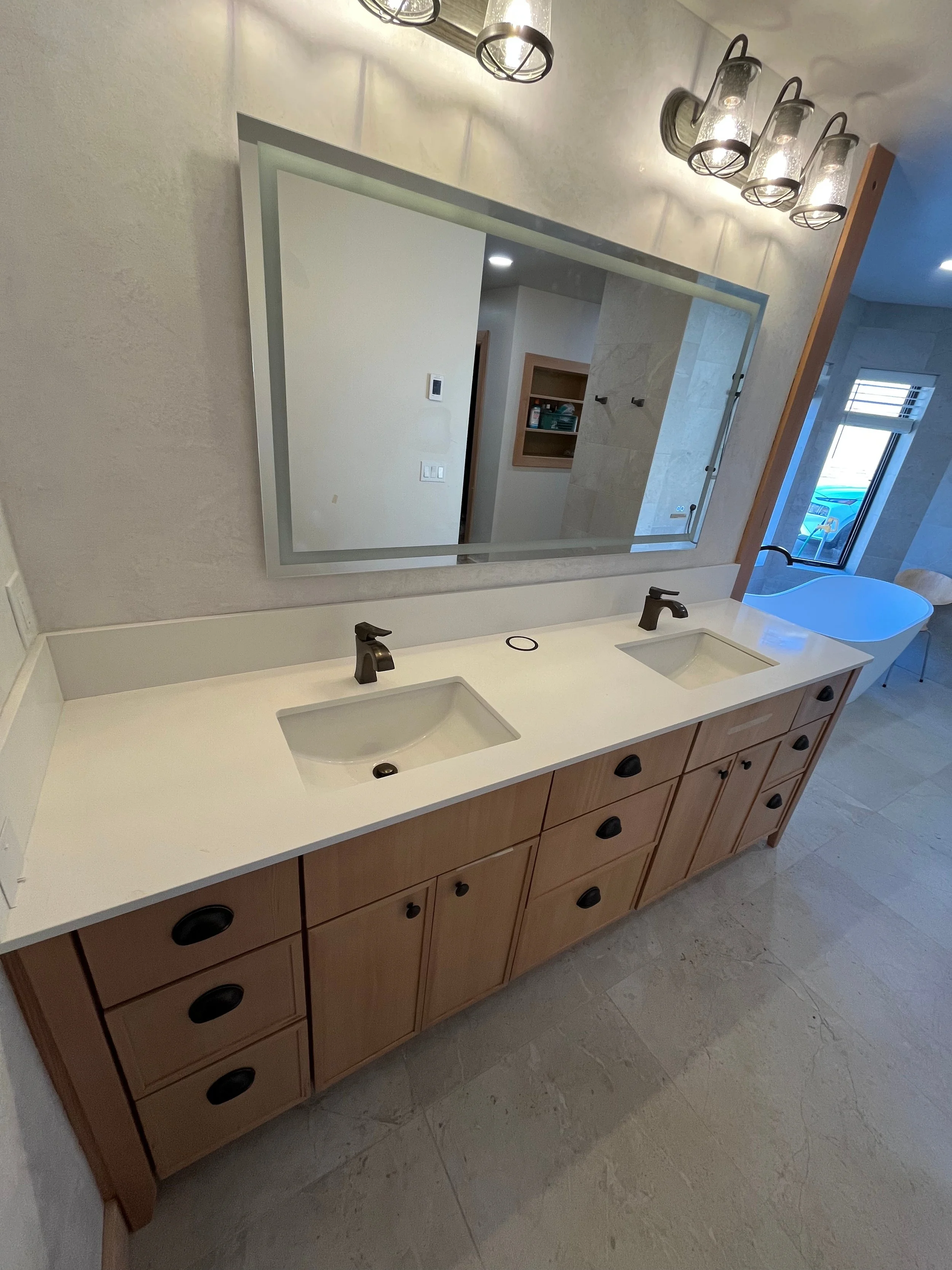 A bathroom vanity with double sinks, a large mirror, and black faucet fixtures. The vanity has wooden cabinetry and is set against a wall painted in a neutral color, with light-colored tiled flooring.