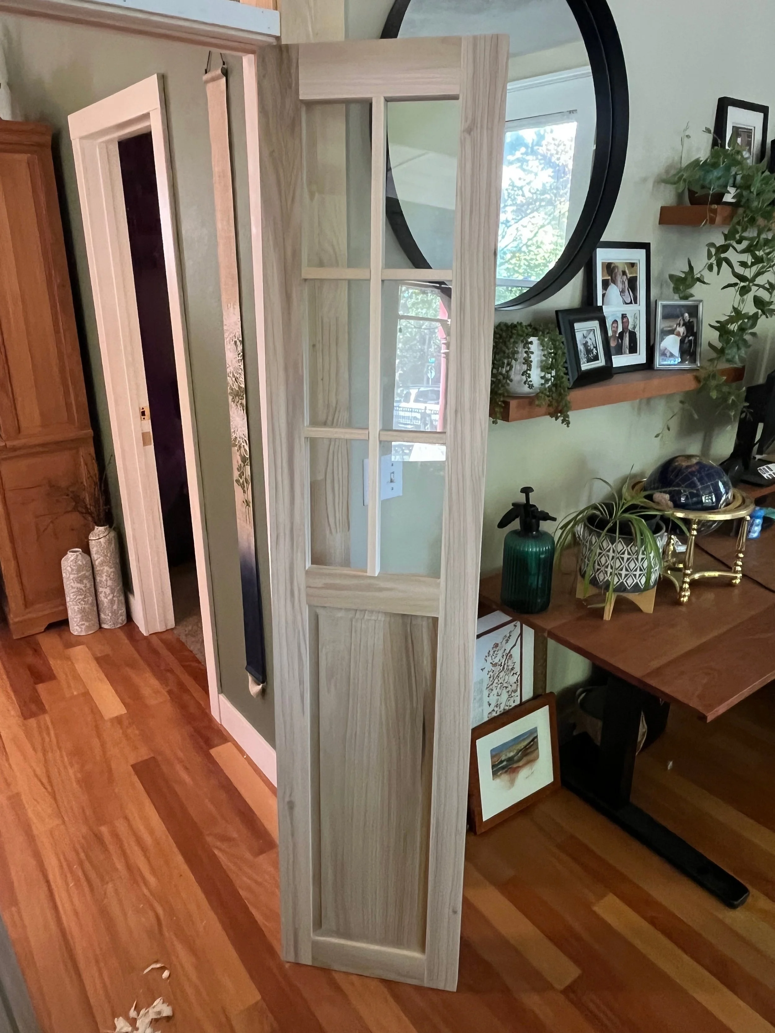 A partially constructed or painted wooden window frame resting on a wooden curb in a room, with a display of framed photos, paintings, and plants on the wall and table nearby.