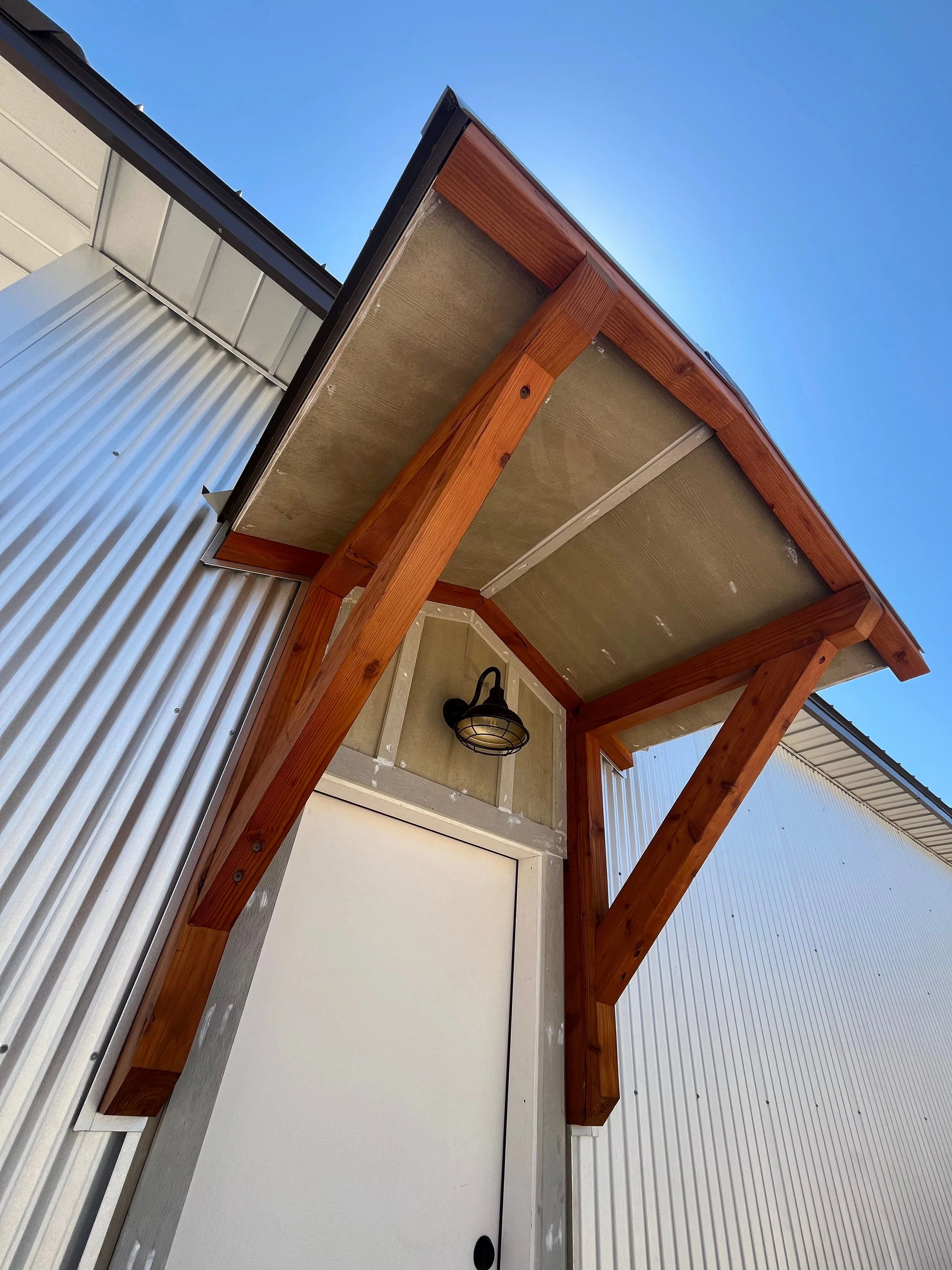 A close-up view of an outdoor patio ceiling with wooden beams, a wall-mounted black lantern, and an overhang with a wooden edge, against a blue sky and metal siding.