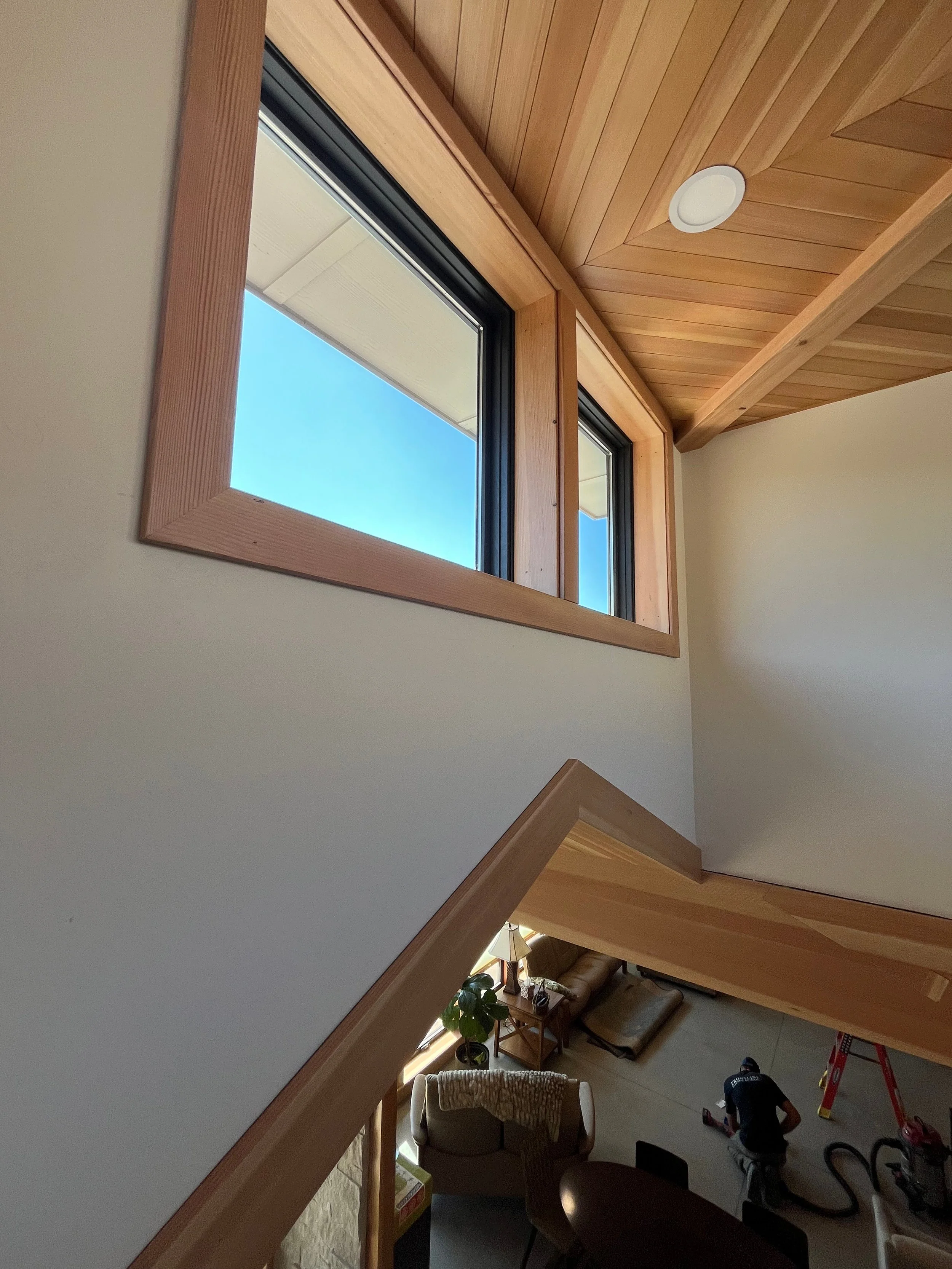 View of a living room with wooden ceiling, window, and a staircase. Visible furniture includes an armchair, side table with a plant, and a person working on the floor with tools and a ladder.