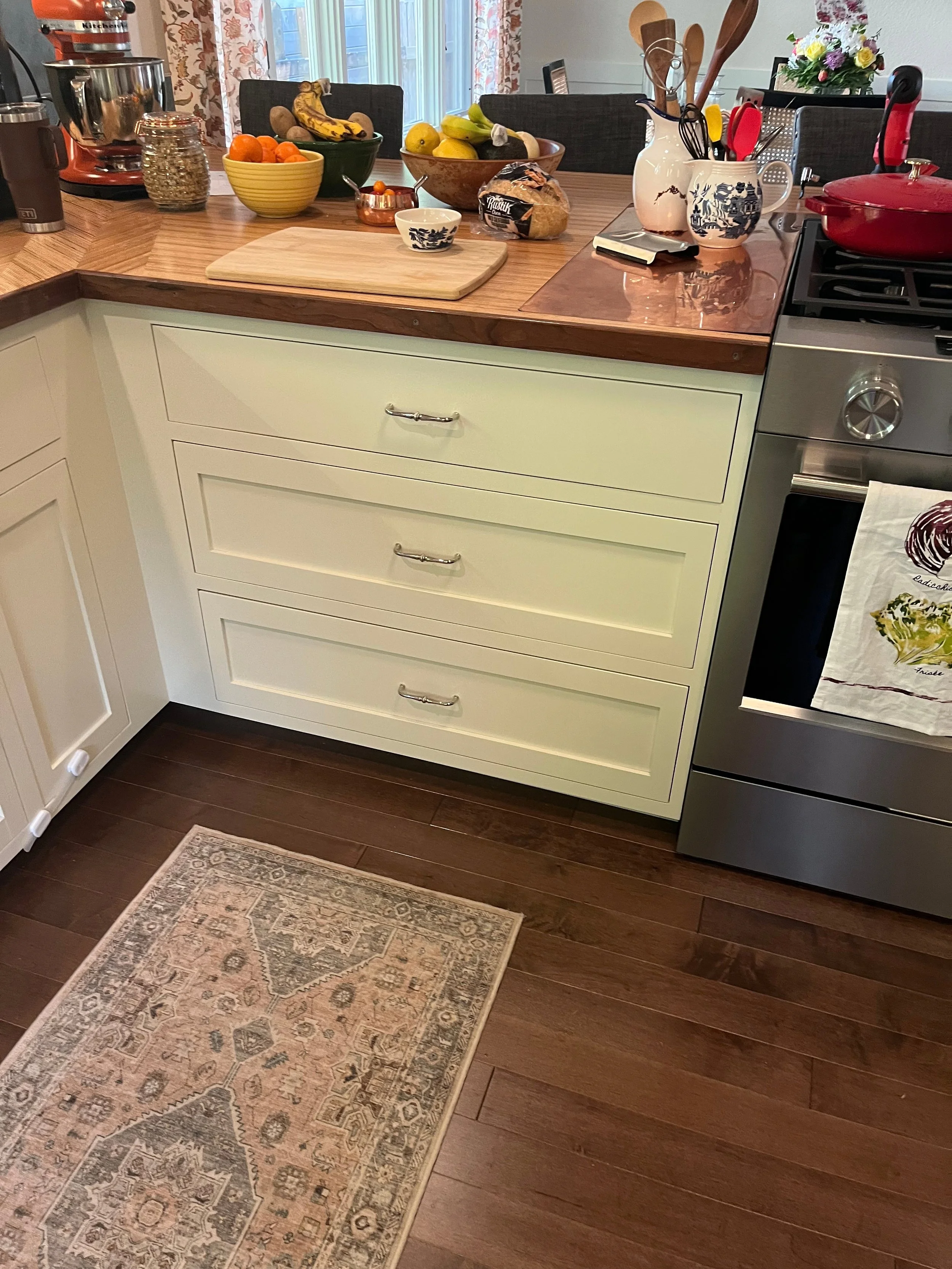 A kitchen counter with various items including a wooden cutting board, bowls of fruit, a bread loaf, a cup, and vases with utensils. There are kitchen appliances like a mixer and stove, with a small rug on a dark wooden floor.