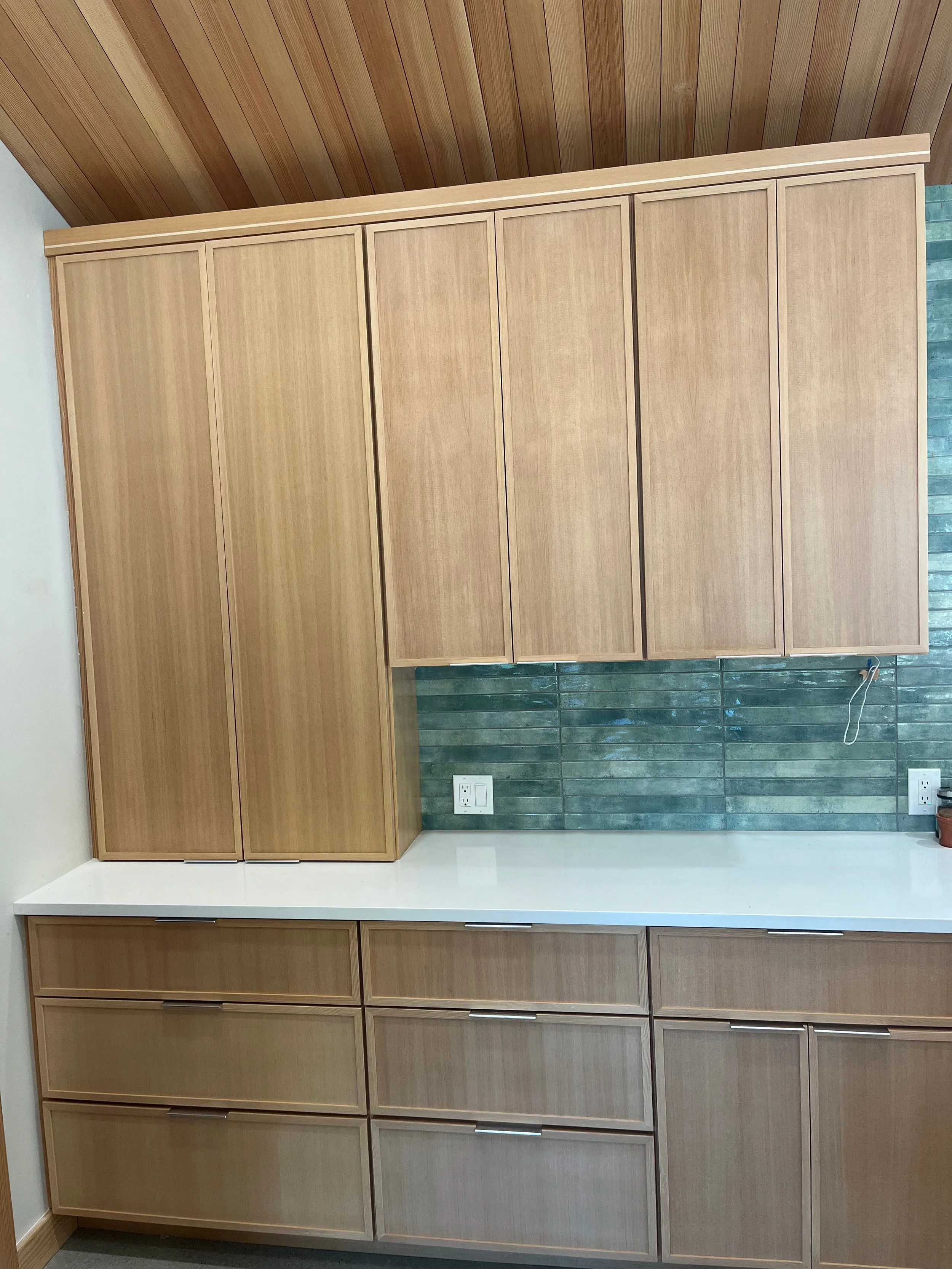 A large setting in a modern kitchen, featuring a row of wooden cabinets with minimalist handles and a white countertop. A green tiled backsplash is visible, along with two electrical outlets and a small potted plant to the right.